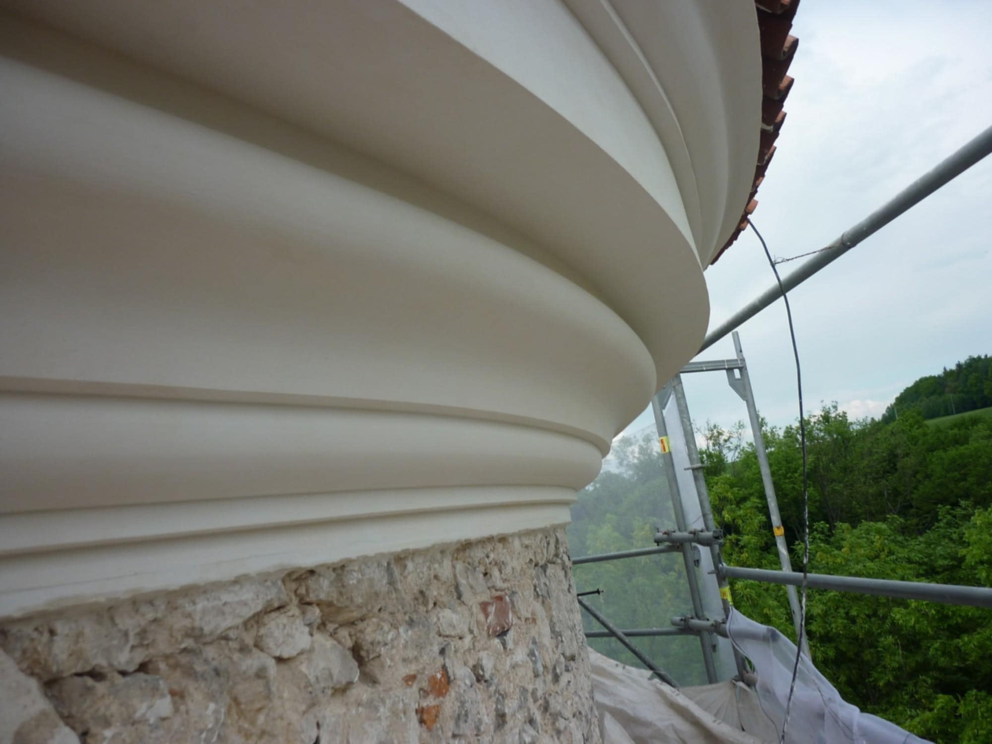 Odnowiona, profilowana elewacja budynku z kamienia i cegły, zabezpieczona rusztowaniem. Widoczne detale architektoniczne i fragment dachu z dachówką.