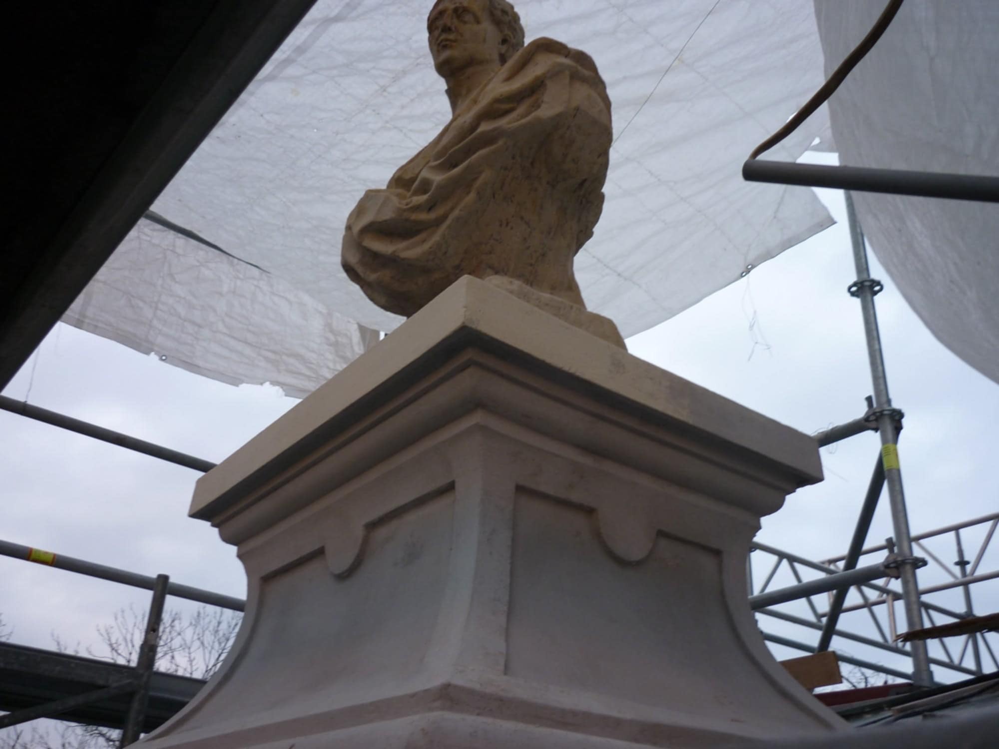Popiersie na postumencie, widoczne rusztowania i osłona. Prace konserwatorskie w toku, ujęcie od dołu, podkreślające monumentalność rzeźby.