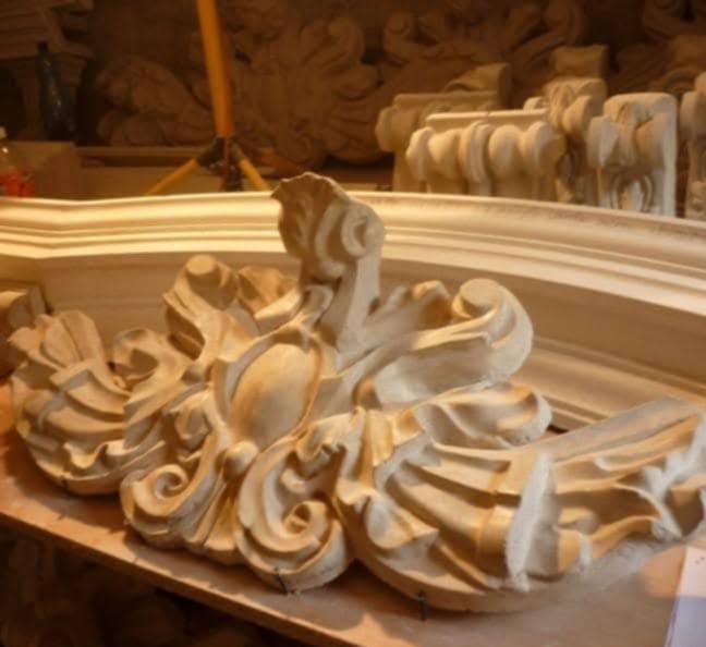 Detal dekoracyjny z jasnego drewna, prawdopodobnie element kominka, z wyraźnymi rzeźbieniami liści i ornamentów, w warsztacie z innymi elementami w tle.