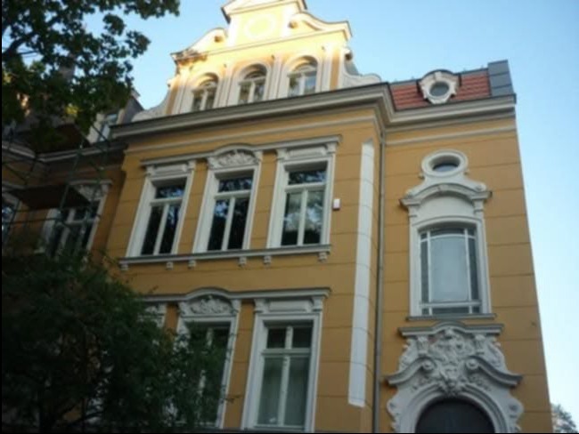 Odrestaurowana fasada kamienicy z żółtą elewacją i białymi zdobieniami okiennymi oraz sztukaterią nad wejściem. Widoczne rusztowania z lewej strony.