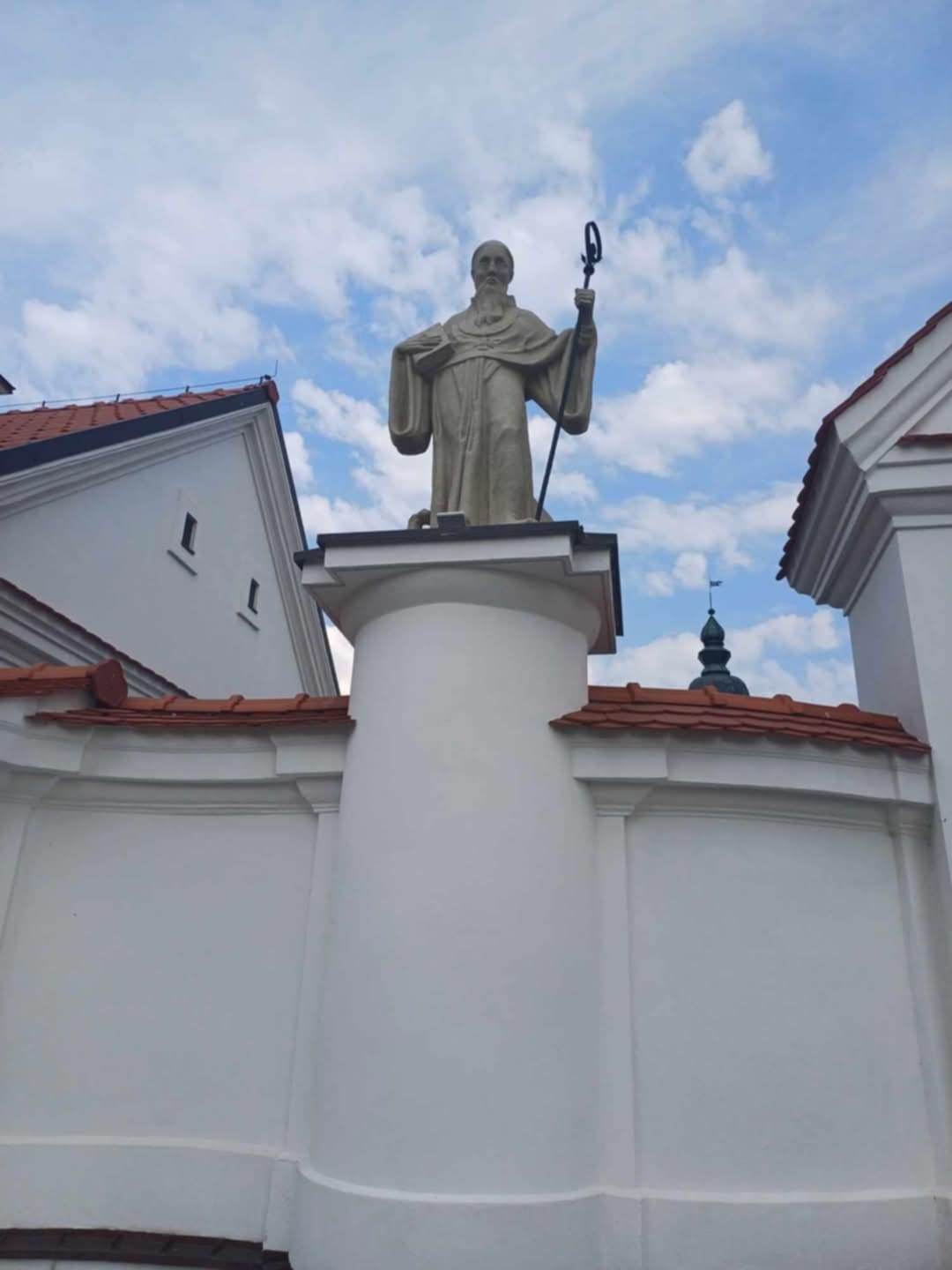 Kamienna figura świętego z laską na kolumnie, na tle białych budynków z czerwoną dachówką i błękitnego nieba z chmurami. Widok z dołu.