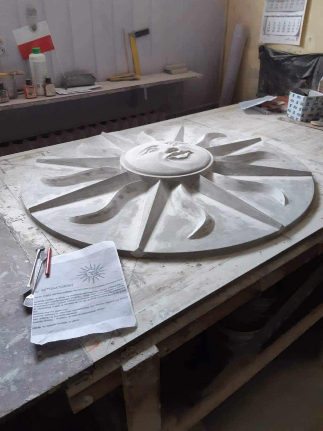 Gipsowy element dekoracyjny w kształcie słońca z twarzą, leżący na roboczym stole w warsztacie. Widoczny dokument z logo 'A. Sztukatorski'.