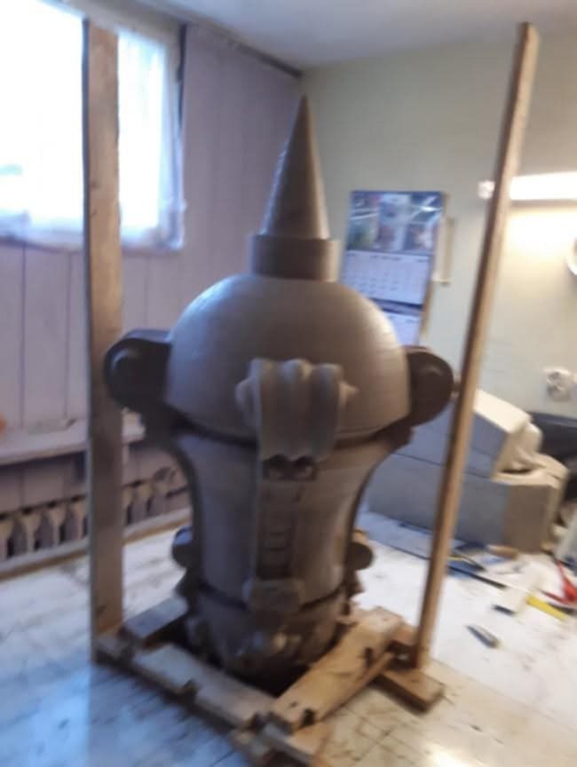 Szara, gliniana forma kominka z dekoracyjnymi elementami, zabezpieczona drewnianą konstrukcją, w jasnym pomieszczeniu z narzędziami na stole.