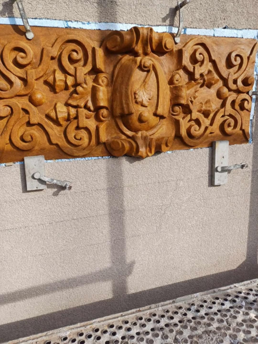 Montaż dekoracyjnego, rzeźbionego elementu fasady w kolorze terakoty, wspartego na metalowych uchwytach, na tle beżowej ściany z fakturą baranka.