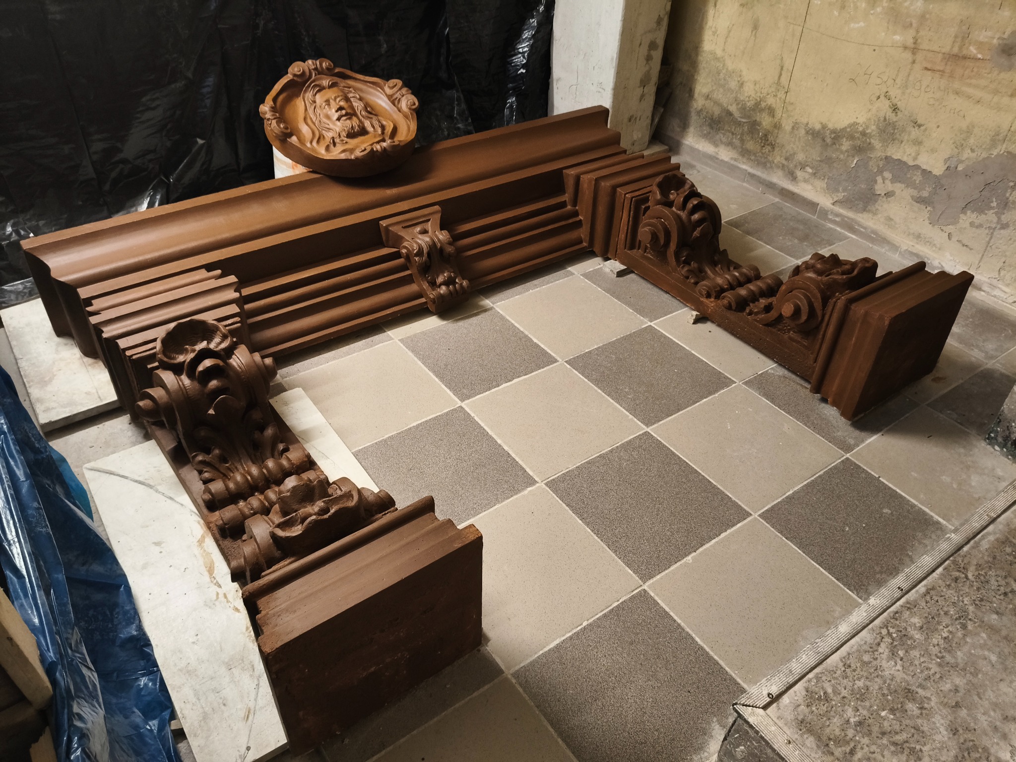 Zdemontowany, odnowiony portal kominkowy z rzeźbionymi elementami, leżący na podłodze w szachownicę. Prace renowacyjne w Gdańsku.