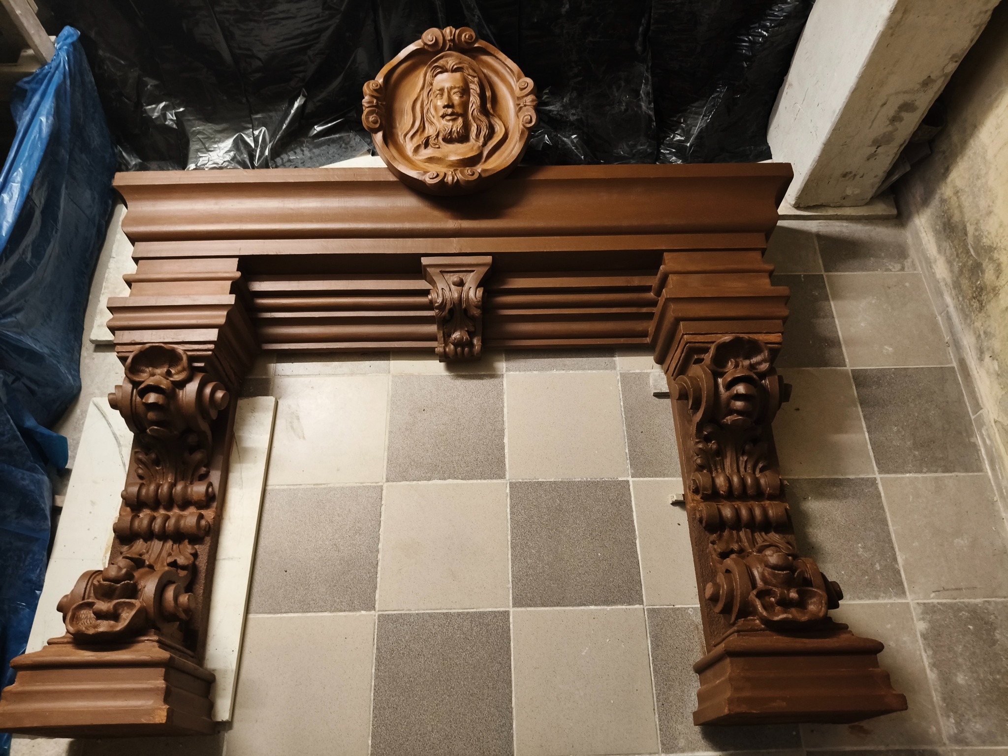 Dekoracyjny, drewniany portal kominkowy z rzeźbioną płaskorzeźbą twarzy, ustawiony na podłodze z płytek w odcieniach szarości, pod ścianą w trakcie prac remontowych.