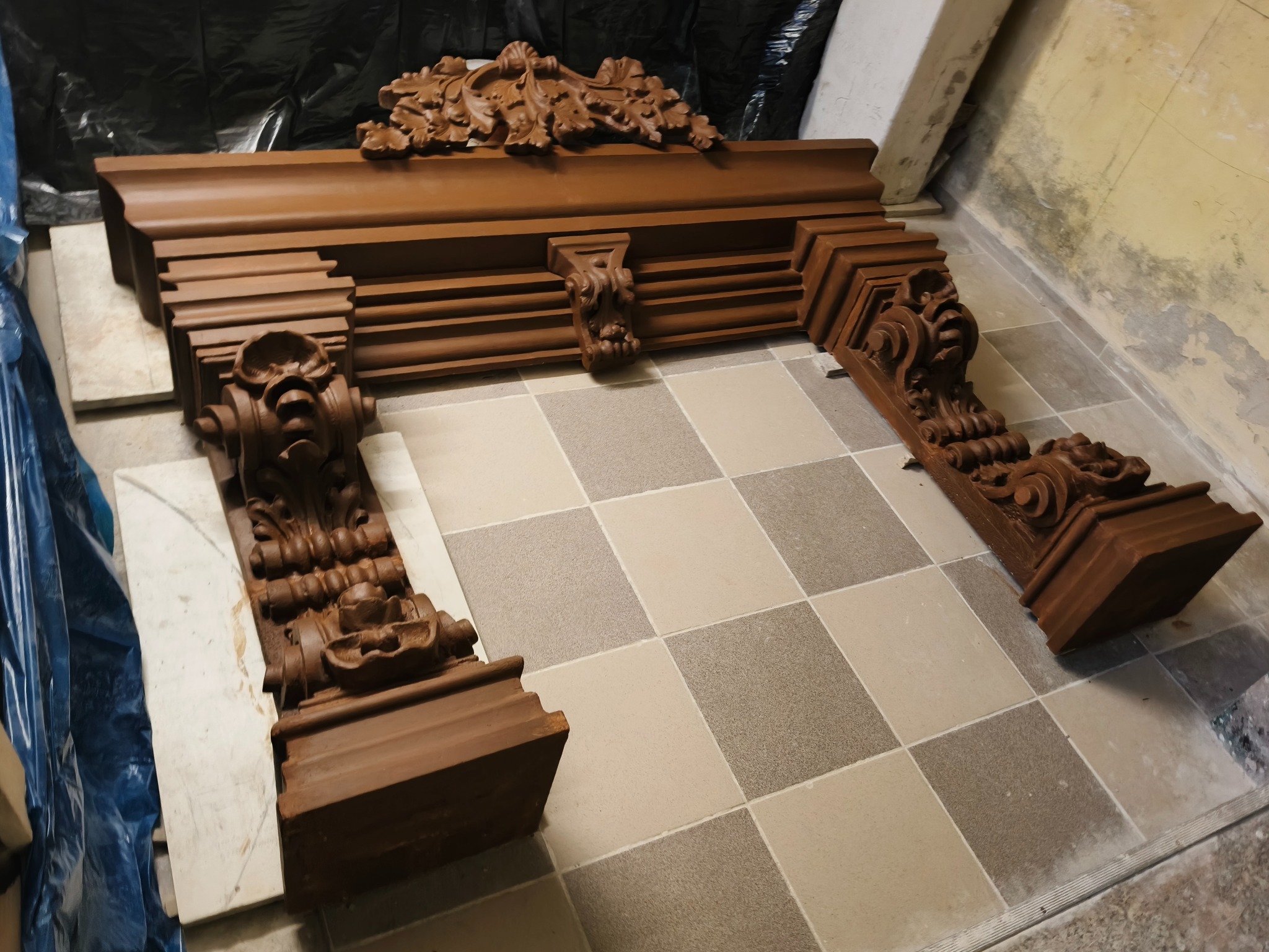 Drewniany, rzeźbiony portal kominkowy w odcieniach brązu z dekoracyjnymi elementami, leżący na podłodze wyłożonej kwadratowymi płytkami.