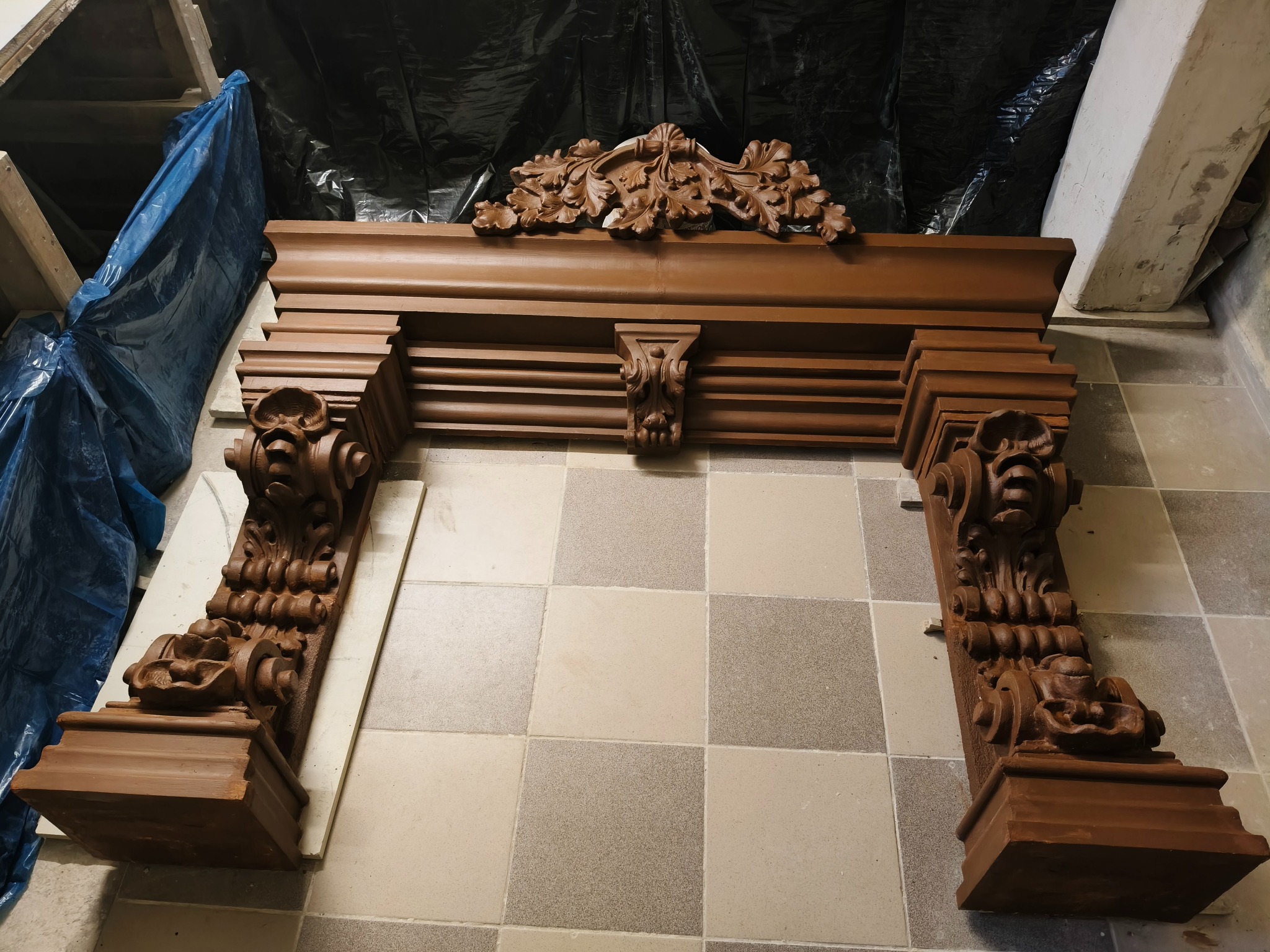 Odrestaurowany, brązowy portal kominkowy z rzeźbionymi elementami roślinnymi i motywami twarzy, ustawiony na podłodze z kwadratowych płytek.