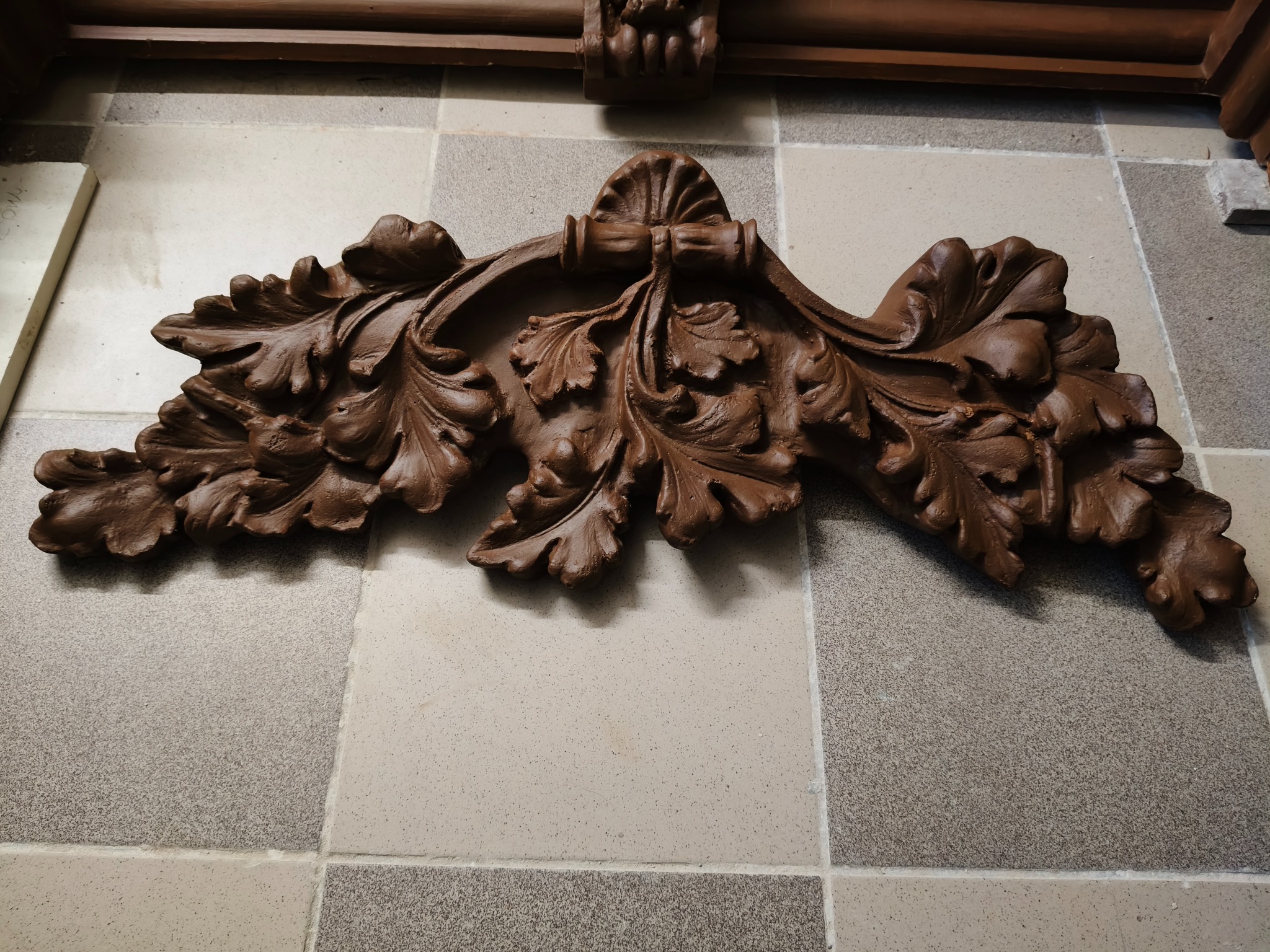 Detal dekoracyjny z brązowego drewna, prawdopodobnie fragment kominka lub pieca kaflowego, na tle wzorzystych płytek. Widoczne elementy rzeźbione w kształcie liści.