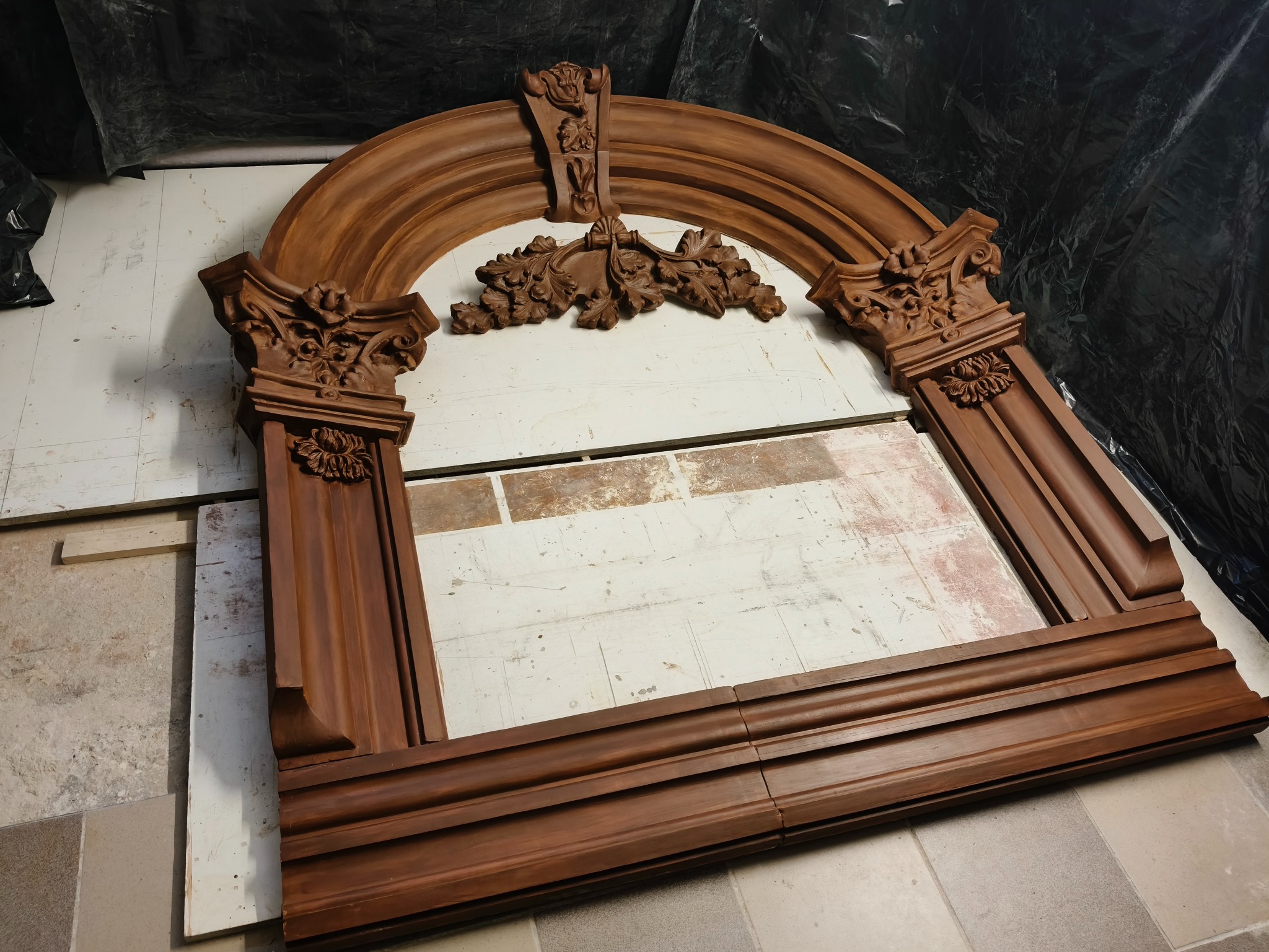 Drewniana, brązowa rama kominka z kolumnami i dekoracyjnymi płaskorzeźbami, leżąca na podłodze w trakcie renowacji. Widoczne ślady farby i niedoskonałości.