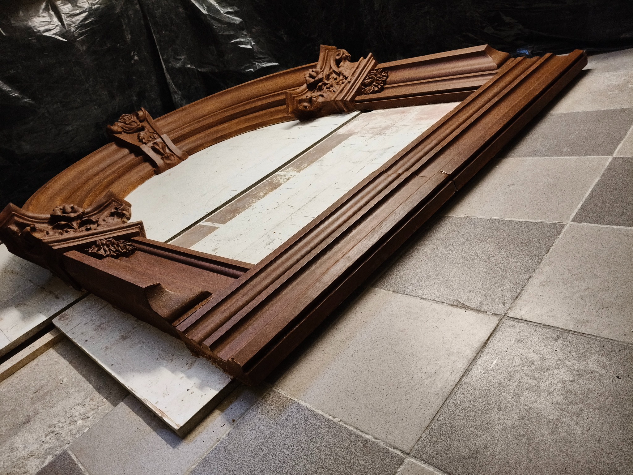 Fragment odrestaurowanej, drewnianej ramy kominka z dekoracyjnymi zdobieniami, leżącej ukośnie na podłodze z płytek. Widoczne detale rzeźbienia i odnowiona powierzchnia drewna.