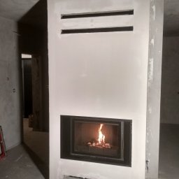 Polmir Łukasz Bartnik - Nowoczesny kominek w surowym wnętrzu. Widoczne palenisko z płomieniami, obudowa w trakcie wykończenia, surowy beton na ścianach i podłodze. Minimalistyczny design.