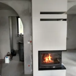 Kominek z płomieniami w surowym wnętrzu, obudowa w trakcie wykończenia, widoczne niedoskonałości ścian i podłogi, co podkreśla etap prac.