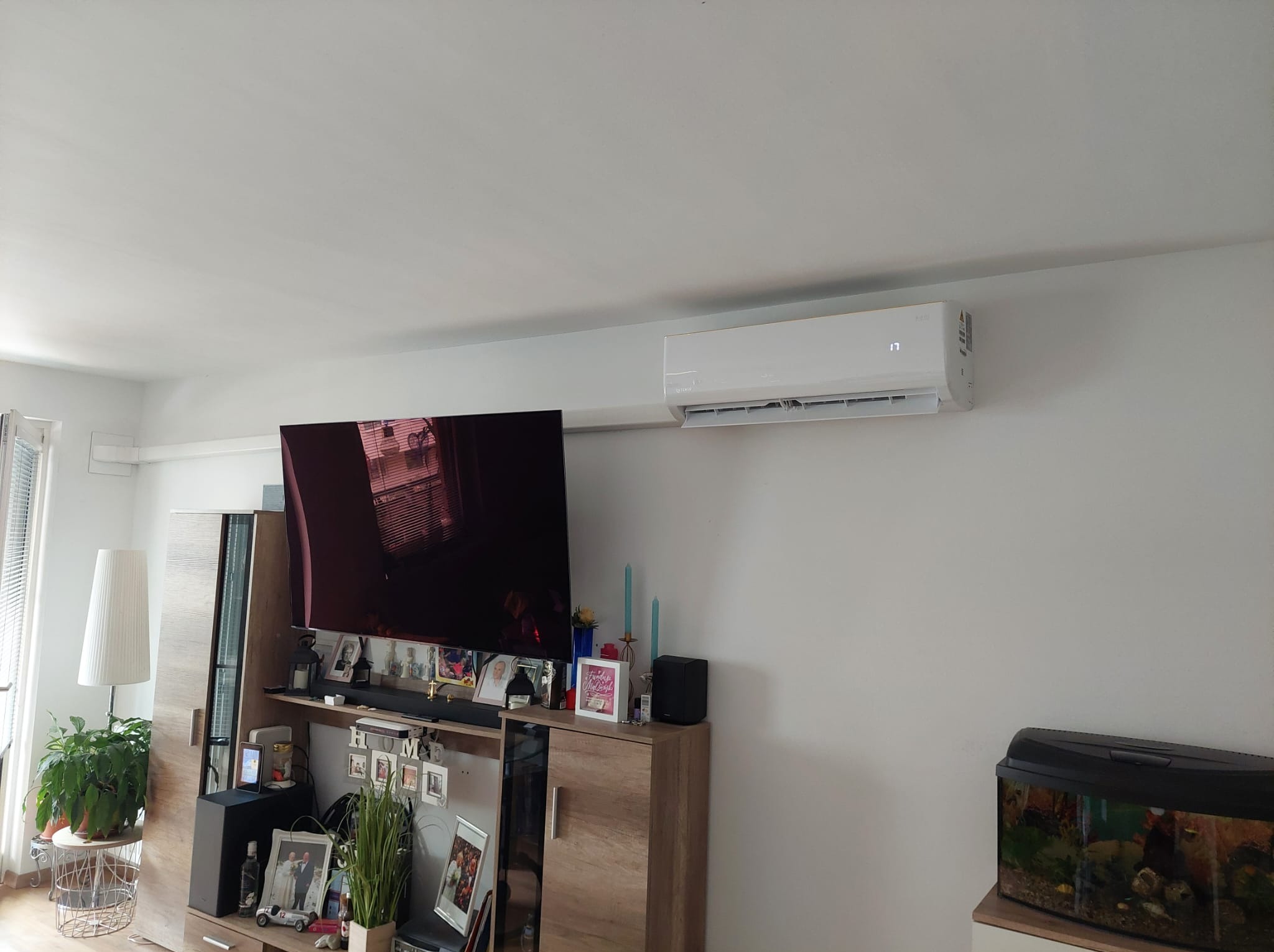 Biała klimatyzacja ścienna w salonie z akwarium i meblami. Widoczny wyświetlacz temperatury na urządzeniu. Nowoczesny design i białe ściany.