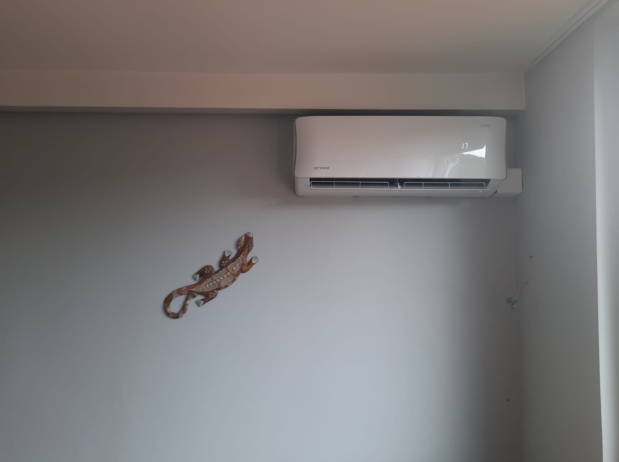 Biała klimatyzacja Rotenso na szarej ścianie w pokoju. Dekoracja ścienna w kształcie jaszczurki. Widoczny wyświetlacz temperatury.