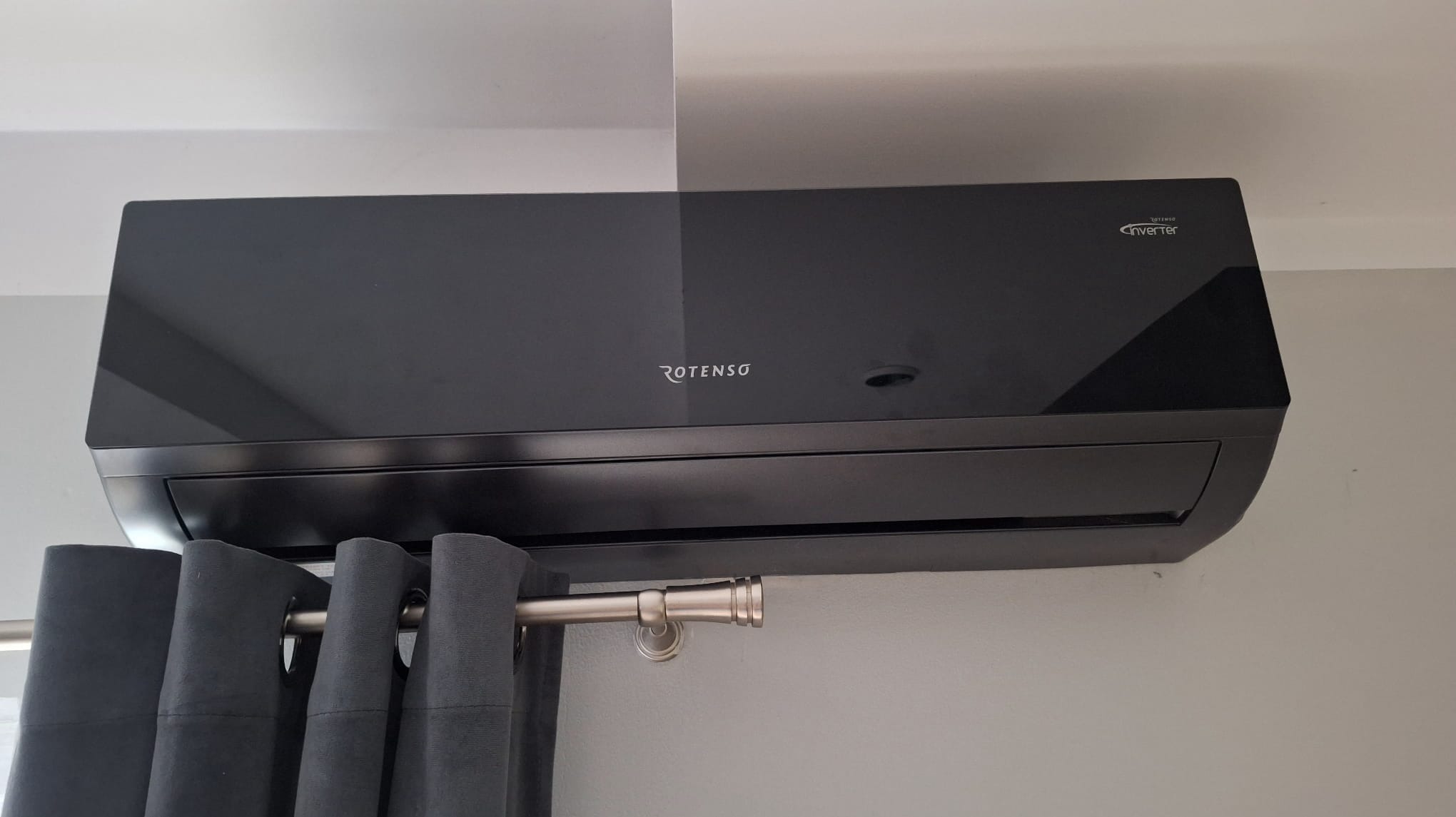 Czarny klimatyzator Rotenso Inverter, zamontowany na ścianie, z zasłonami w tle. Elegancki design i nowoczesne rozwiązanie chłodzenia.