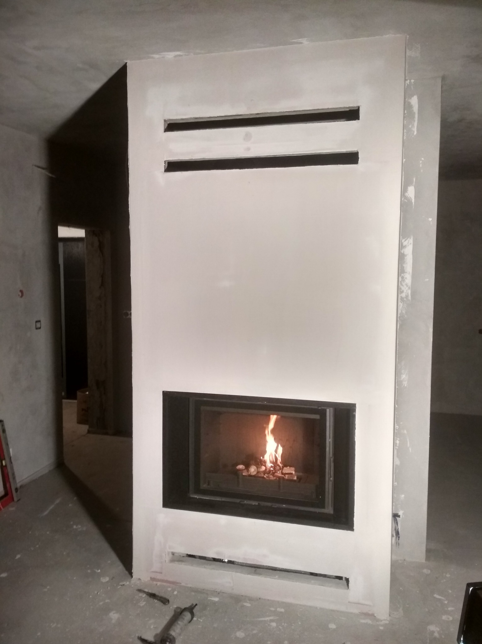 Nowoczesny kominek w surowym wnętrzu. Widoczne palenisko z płomieniami, obudowa w trakcie wykończenia, surowy beton na ścianach i podłodze. Minimalistyczny design.