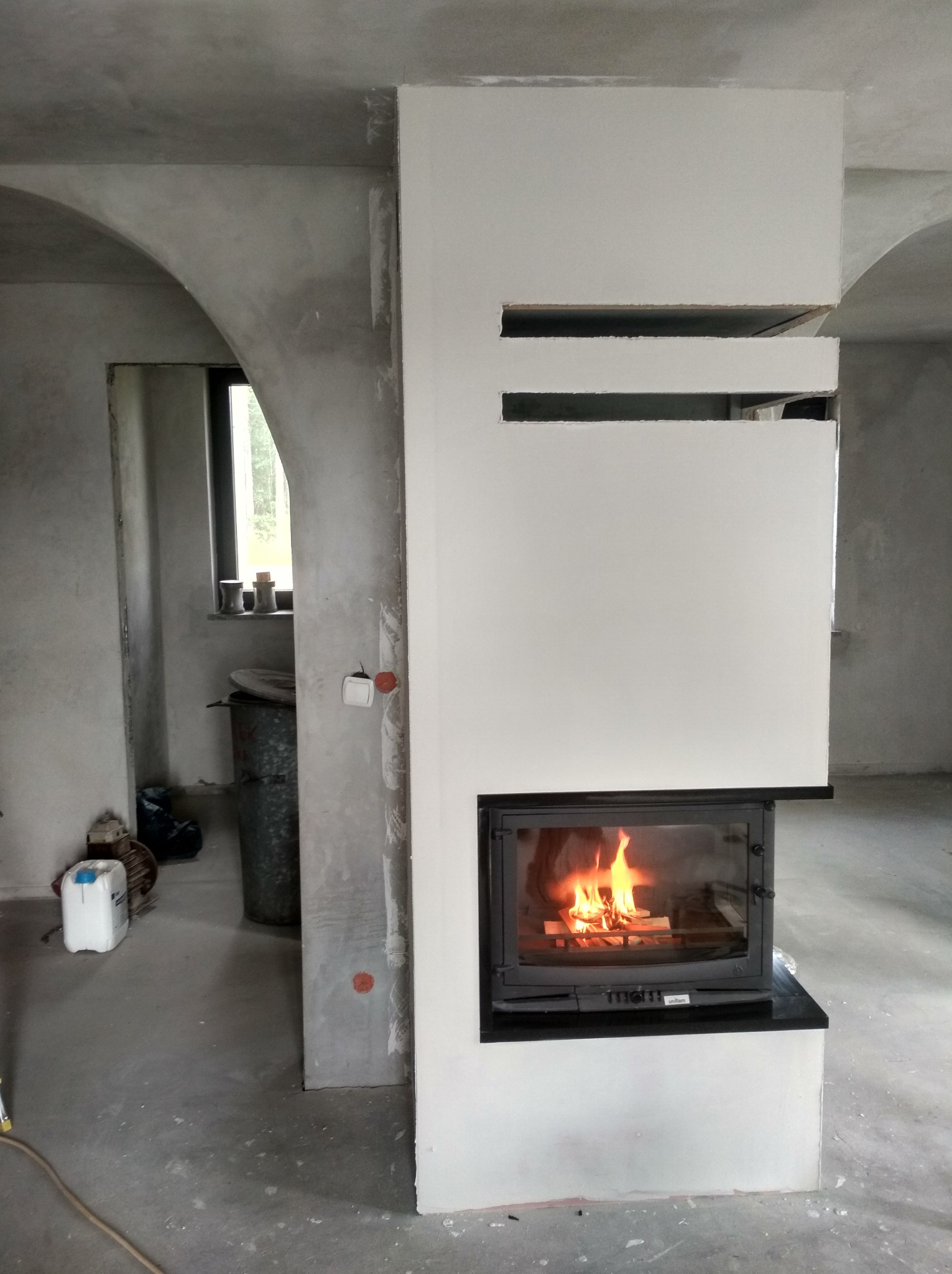 Kominek z płomieniami w surowym wnętrzu, obudowa w trakcie wykończenia, widoczne niedoskonałości ścian i podłogi, co podkreśla etap prac.