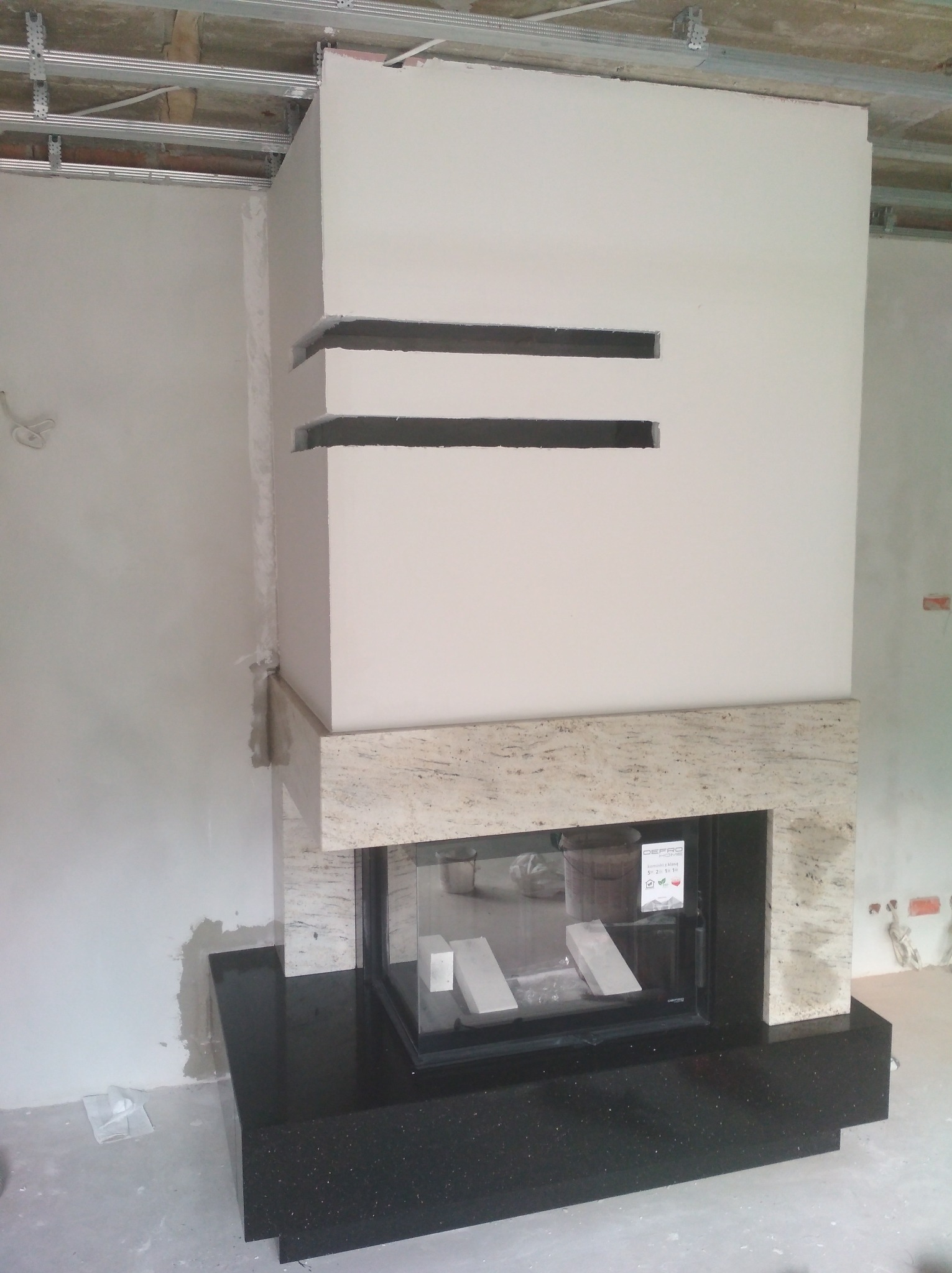 Nowoczesny kominek w trakcie montażu, z obudową z jasnego kamienia i czarnym blatem, widoczne elementy konstrukcyjne sufitu podwieszanego.