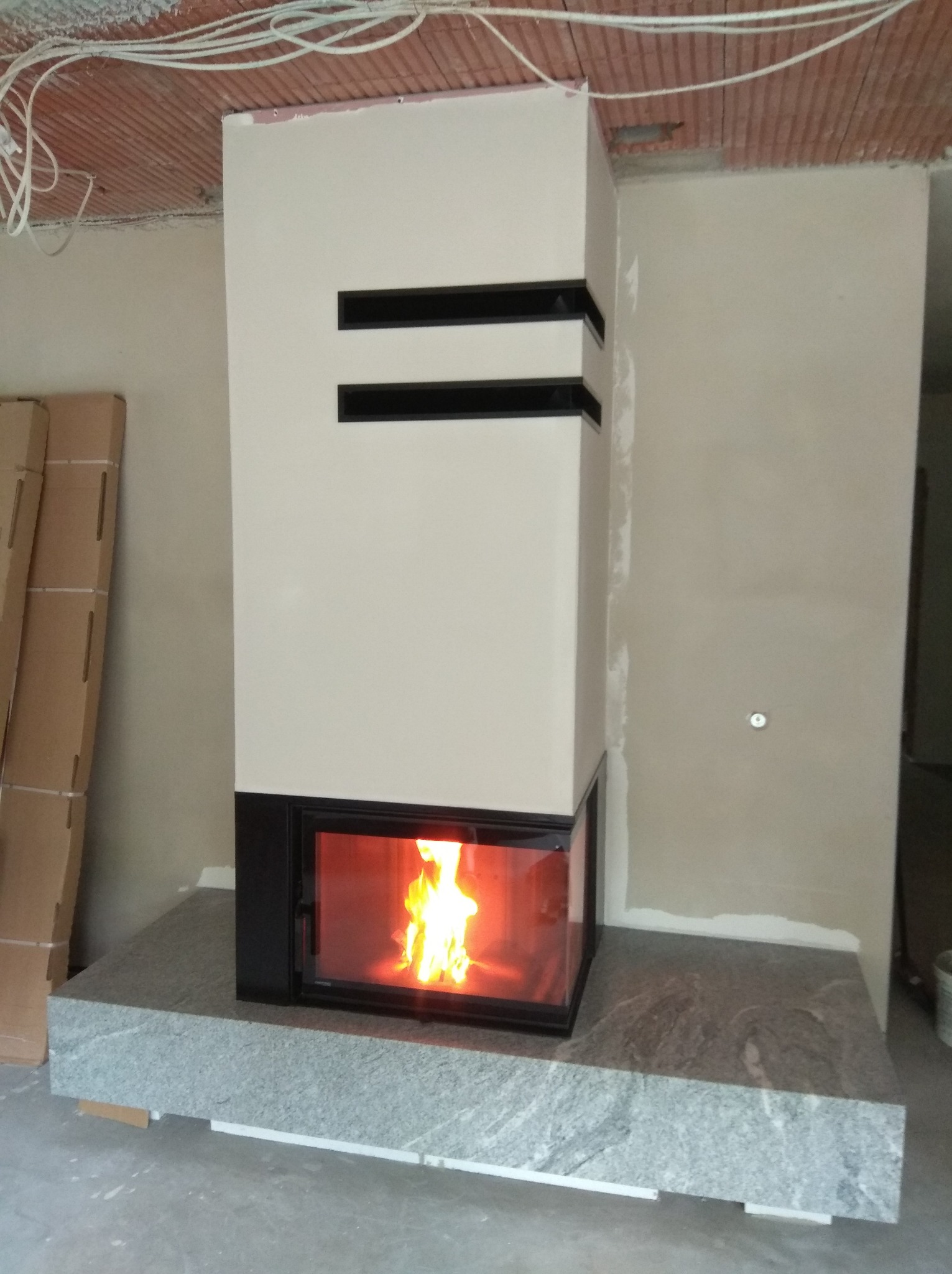 Nowoczesny kominek z czarną obudową i płomieniami, umieszczony na granitowej płycie w surowym wnętrzu. Widoczne elementy instalacji na suficie.