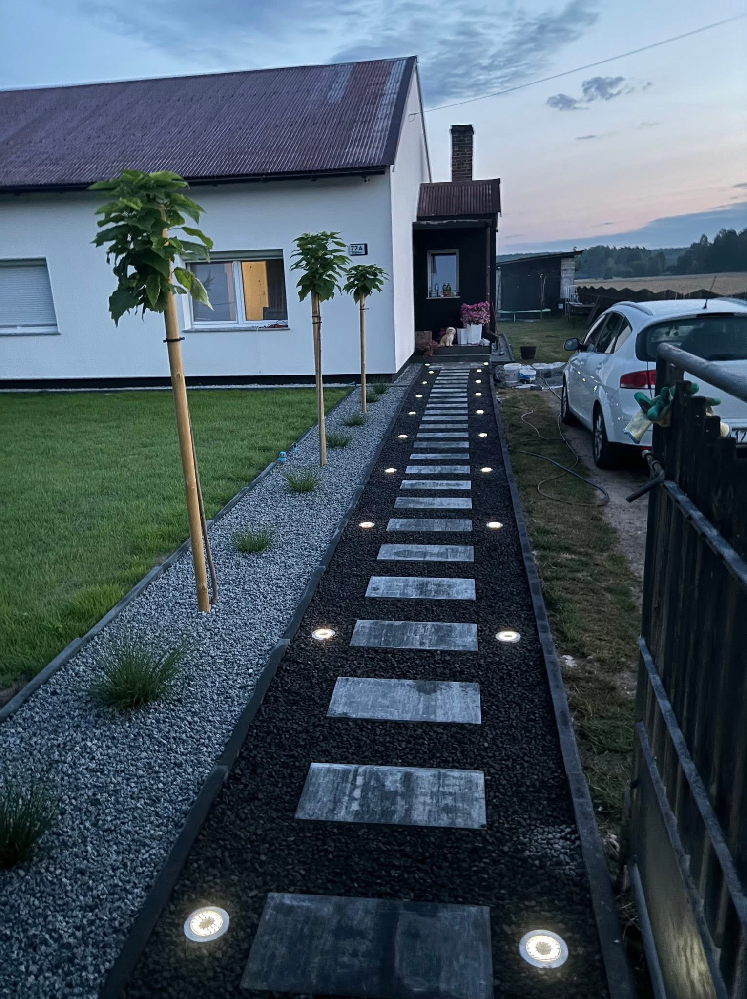 Oświetlona ścieżka z płyt prowadząca do domu, obok trawnik i drzewka. Dekoracyjny żwir i lampy podkreślają estetykę ogrodu po zmroku.