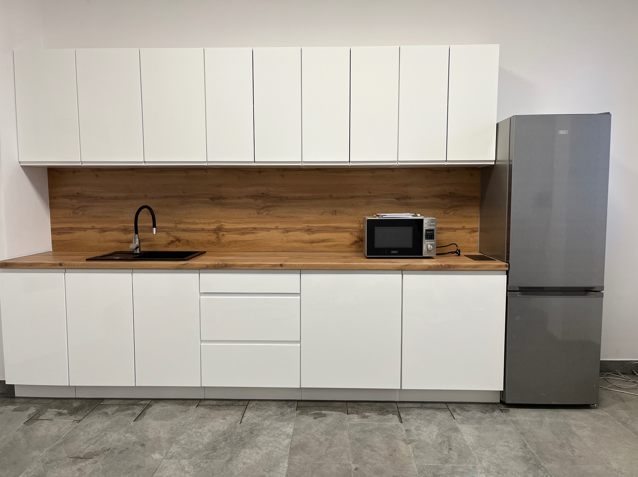 Nowoczesna kuchnia z białymi szafkami, drewnianym blatem i szarą lodówką. Minimalistyczny design z czarną armaturą i mikrofalówką.