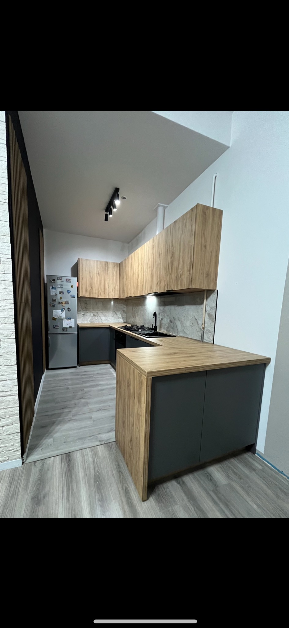 Nowoczesna kuchnia z frontami w odcieniach drewna i szarości, marmurowy blat, czarna armatura i oświetlenie punktowe. Kontrastujące panele ścienne.