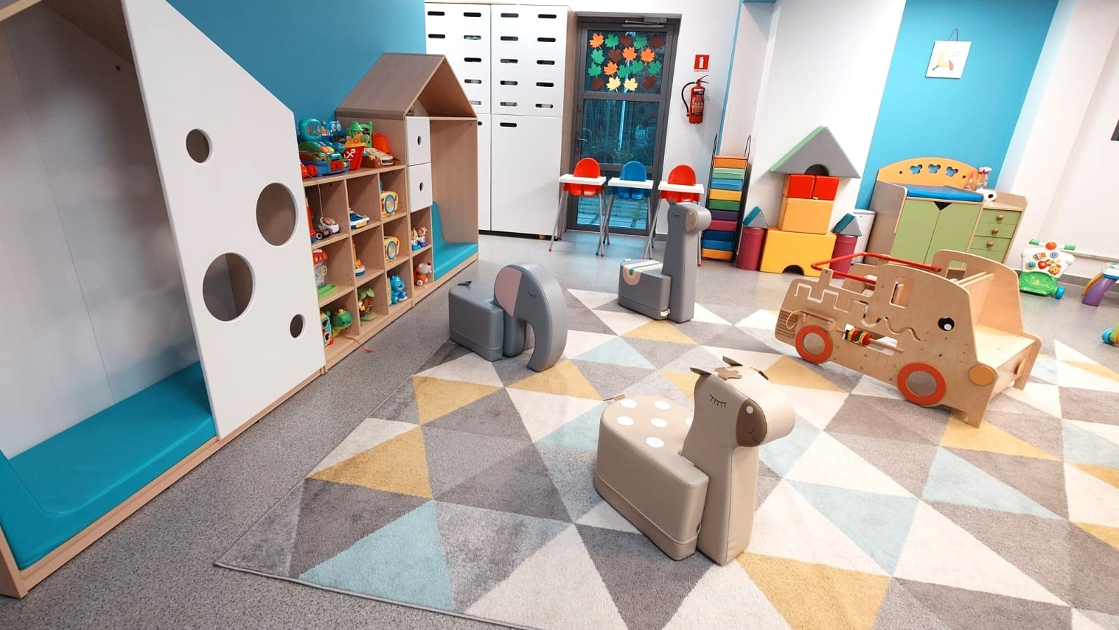 Kolorowa sala zabaw w żłobku: drewniane meble-zabawki (słoń, żyrafa, auto), regały z zabawkami, krzesełka do karmienia, dywan w geometryczne wzory, niebieska ściana.