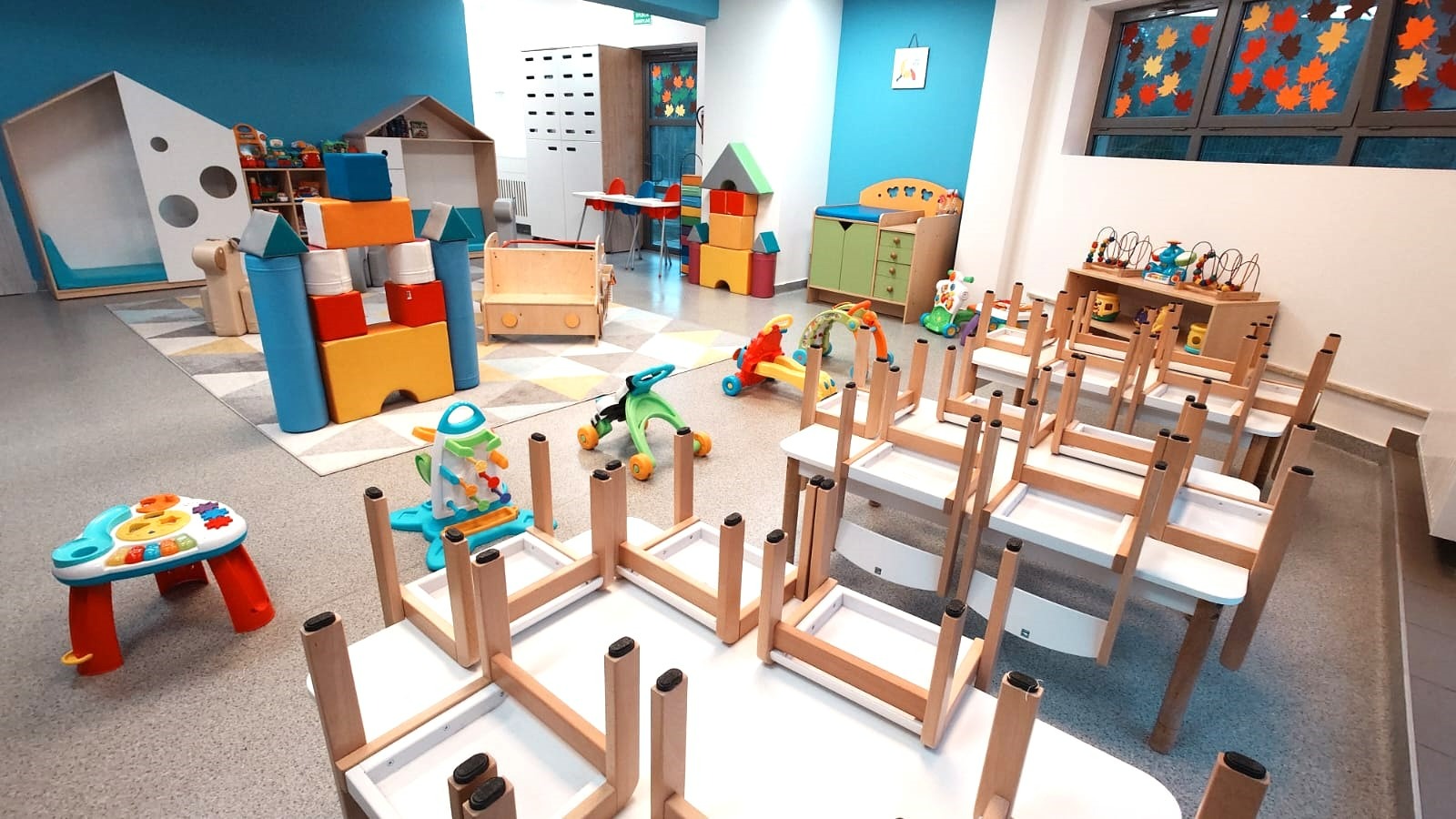 Kolorowa sala zabaw dla dzieci z odwróconymi krzesłami na stołach, zabawki edukacyjne i dekoracje okienne z motywem liści.