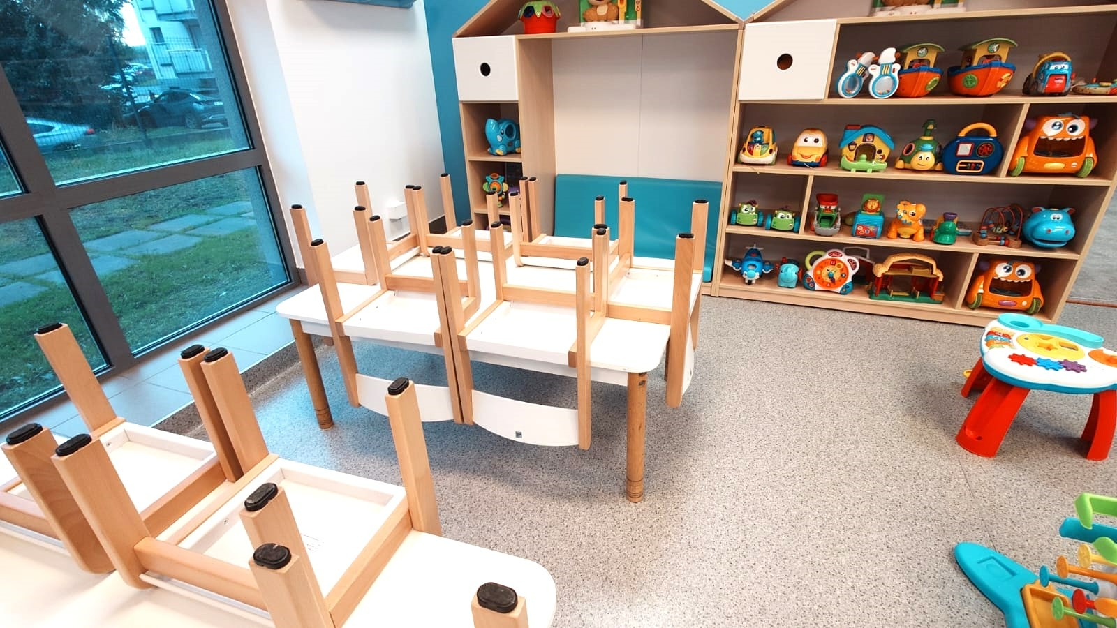 Sala zabaw w przedszkolu: białe stoliki z krzesłami ustawionymi do góry nogami, regał z zabawkami w tle, okno z widokiem na zewnątrz, jasne wnętrze.