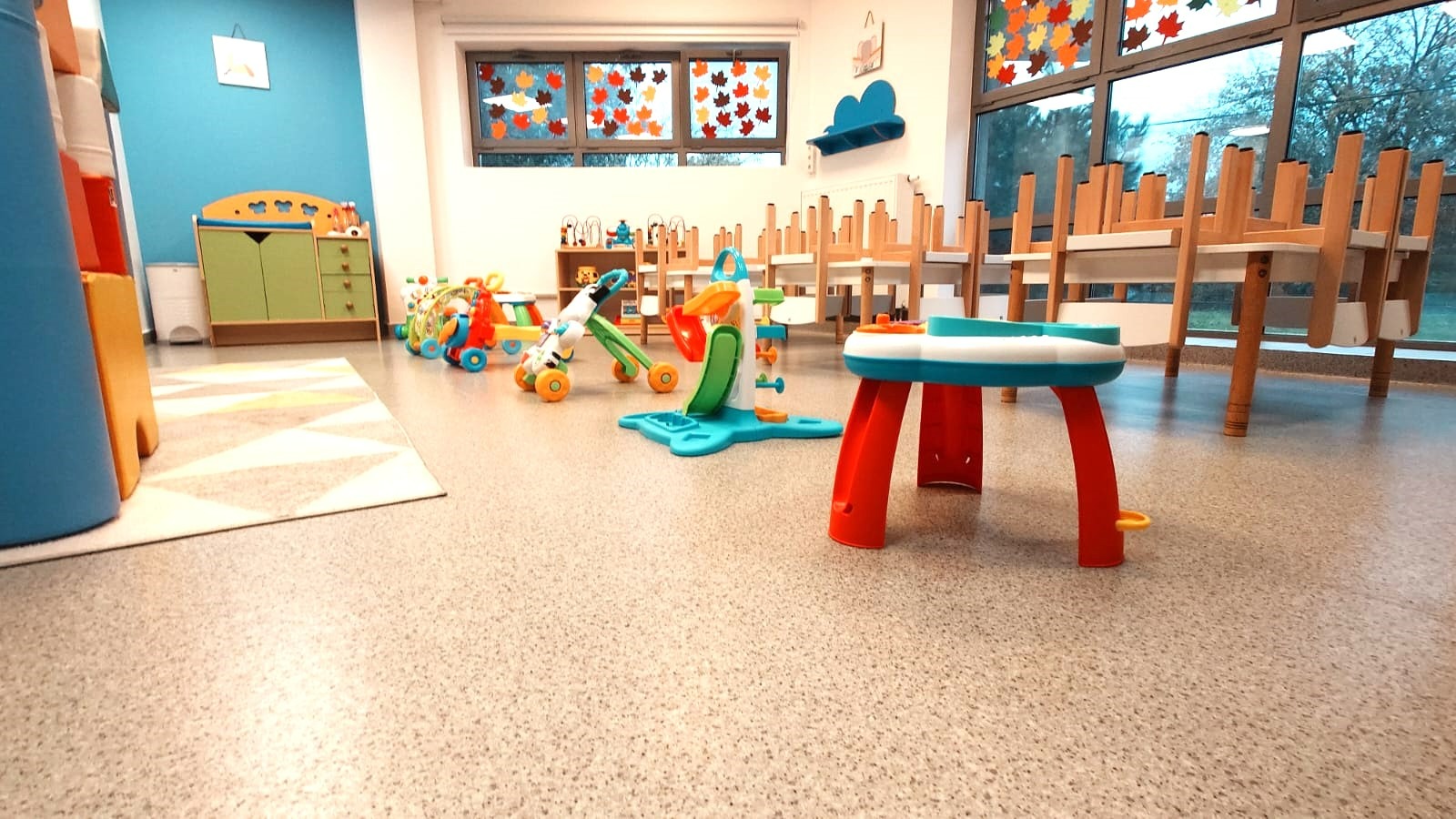 Sala zabaw w żłobku z kolorowymi zabawkami, niskimi mebelkami i dekoracjami okiennymi z motywem liści. Krzesła ułożone na stołach.