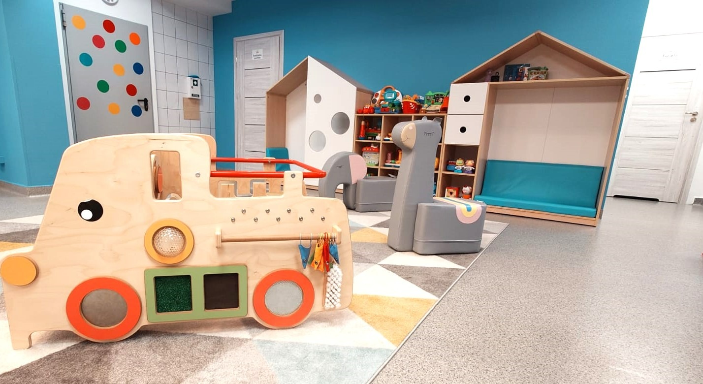 Kolorowa sala zabaw z drewnianą lokomotywą sensoryczną, regałami w kształcie domków i krzesełkami-zwierzątkami na wzorzystym dywanie. Błękitne ściany.