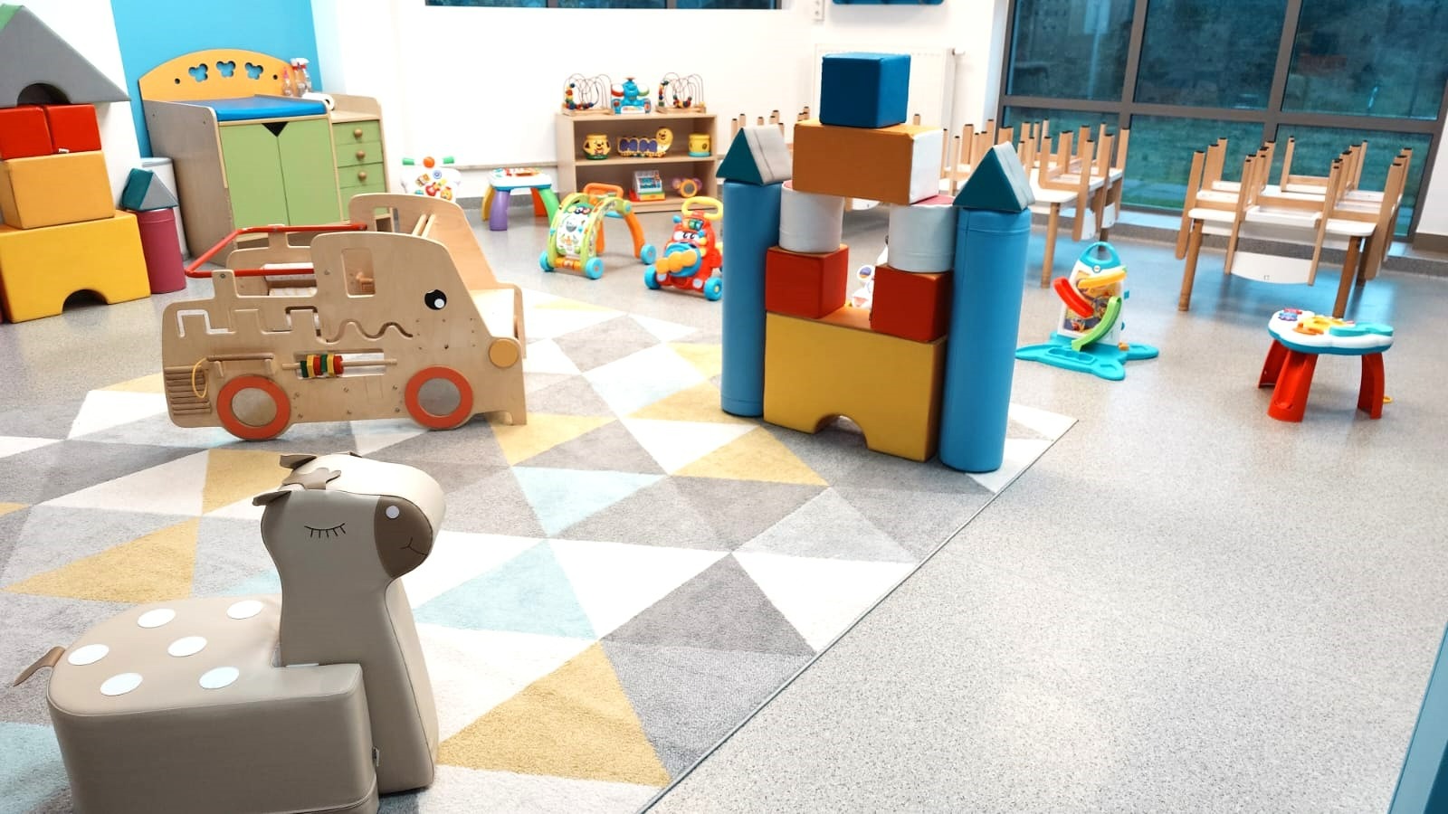 Jasne, przestronne wnętrze sali zabaw z drewnianymi zabawkami, kolorowymi klockami i krzesełkami przy oknie. Podłoga wyłożona dywanem w geometryczne wzory.