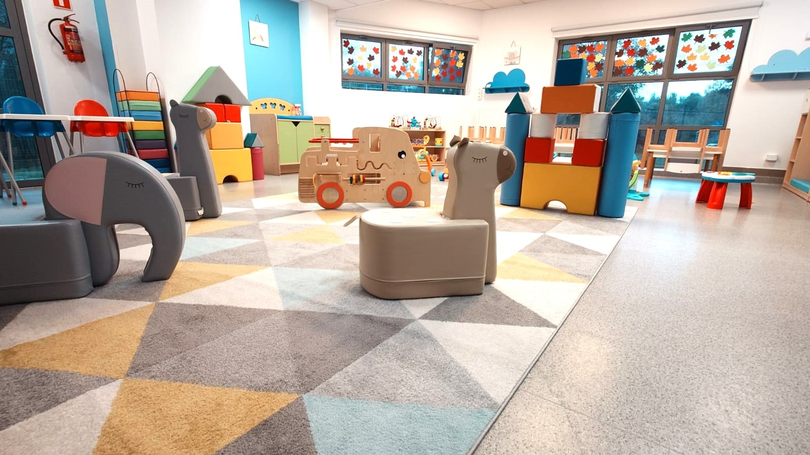 Kolorowa sala zabaw z dywanem w geometryczne wzory, meblami w kształcie zwierząt i drewnianą lokomotywą. Okna ozdobione jesiennymi liśćmi.