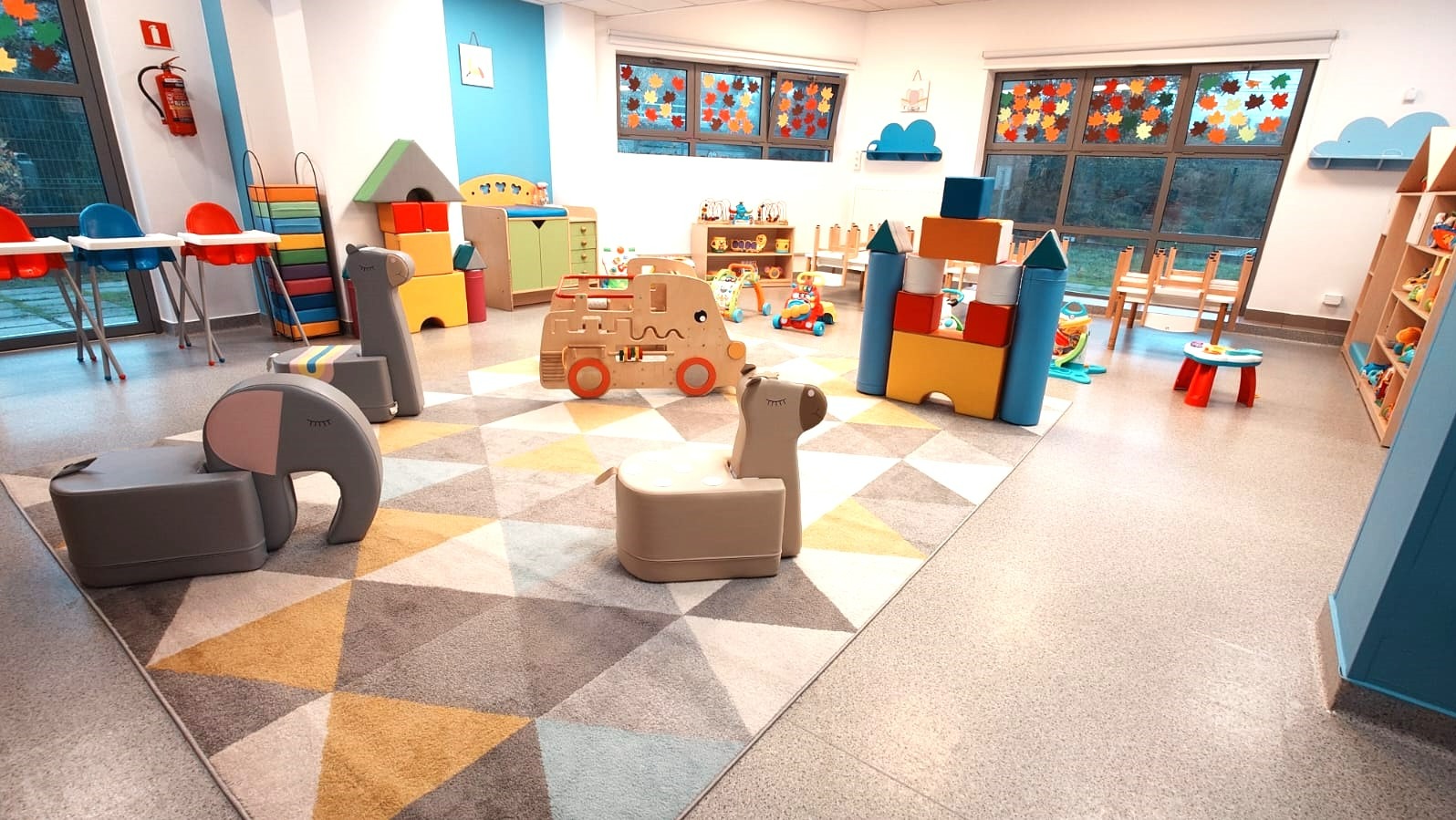 Kolorowa sala zabaw z meblami w kształcie zwierząt, drewnianymi zabawkami i dywanem w geometryczne wzory. Okna zdobią jesienne dekoracje.