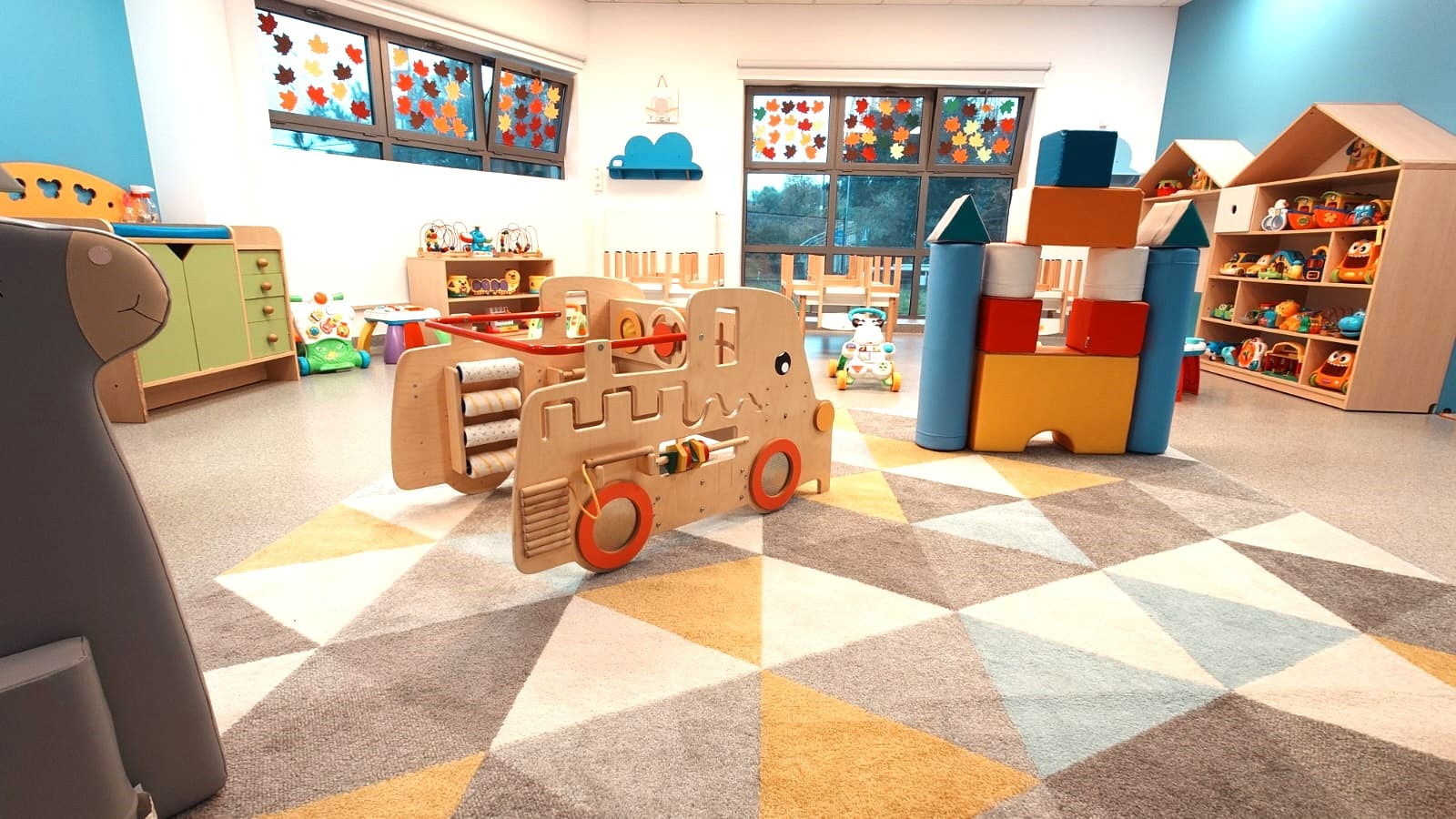 Jasne wnętrze sali zabaw z drewnianym pojazdem edukacyjnym na pierwszym planie, kolorowe klocki i regały z zabawkami w tle, okna ozdobione jesiennymi liśćmi.