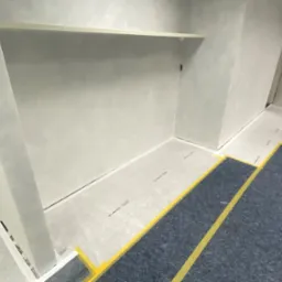 Szara łazienka w trakcie remontu: zabezpieczona podłoga, świeżo położone gładzie na ścianach, widoczny fragment umywalki. W tle drzwi.