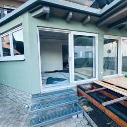 Krzysztof Ubych Usługi Budowlane - Zabudowa tarasu z zieloną elewacją, widoczne drewniane elementy konstrukcyjne i schody z metalowej kraty. Wnętrze w trakcie wykończenia, okna przesuwne.