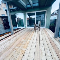 Krzysztof Ubych Usługi Budowlane - Drewniany taras w trakcie budowy przy domu z przesuwnymi drzwiami balkonowymi. Widoczne deski, podkonstrukcja i fragment domu z rynną.