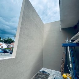 Krzysztof Ubych Usługi Budowlane - Świeżo otynkowany balkon z wykończoną elewacją w neutralnym odcieniu szarości. Widoczne rusztowania i narzędzia malarskie. Tło stanowi pochmurne niebo i zabudowa miejska.