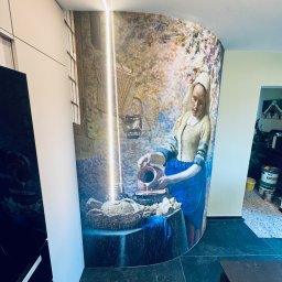 Krzysztof Ubych Usługi Budowlane - Ściana wykończona fototapetą z motywem obrazu Vermeera 'Mleczarka', wkomponowana w nowoczesną zabudowę kuchenną z oświetleniem LED. Ciemna podłoga z płytek.