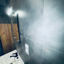 Krzysztof Ubych Usługi Budowlane - Nowoczesna łazienka w Bielawie: szare płytki, biała umywalka z czarną armaturą, lustro z oświetleniem LED i drewniane elementy wykończenia.