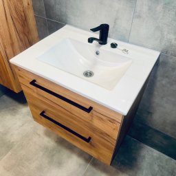 Krzysztof Ubych Usługi Budowlane - Nowoczesna szafka pod umywalkę z drewnianymi frontami i czarnymi uchwytami, biała umywalka i czarna bateria. Szare płytki na ścianie i podłodze.