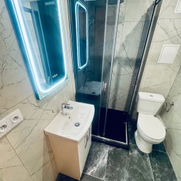 Krzysztof Ubych Usługi Budowlane - Wykończona łazienka z kabiną prysznicową, umywalką z szafką i toaletą. Ściany i podłoga wyłożone płytkami imitującymi marmur. Lustro z podświetleniem LED.