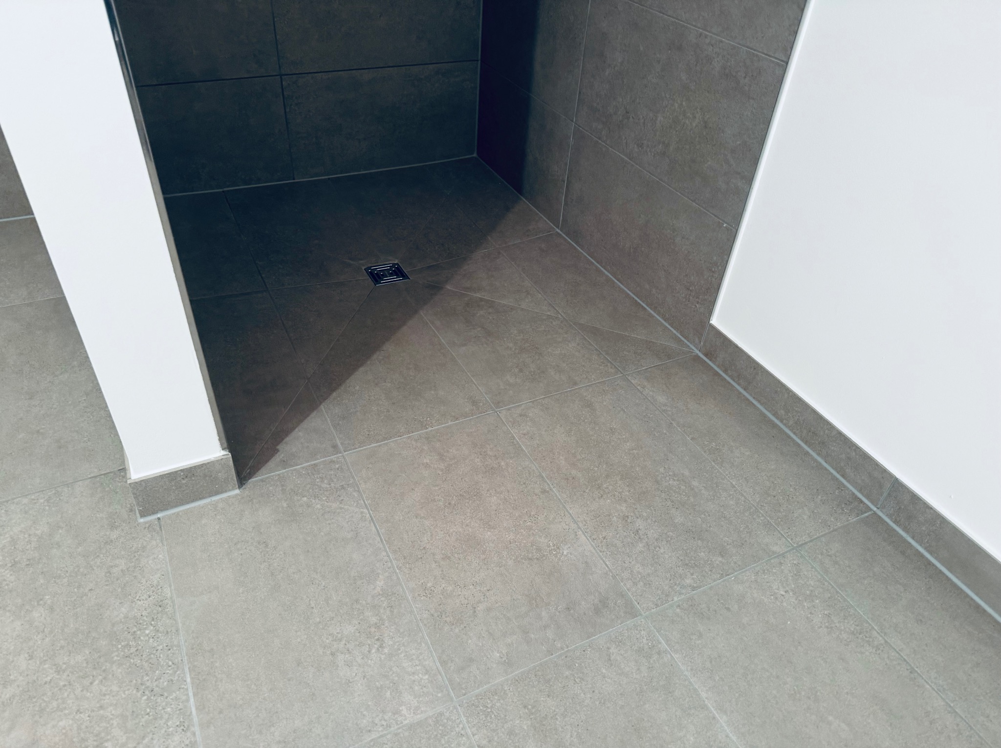 Nowoczesny prysznic z odpływem liniowym i szarą, kwadratową glazurą na ścianach i podłodze. Minimalistyczny design, białe ściany, precyzyjne wykończenie.