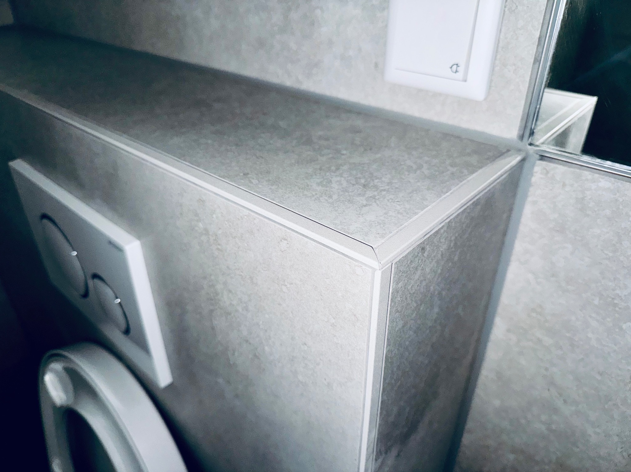 Szary blat z płytek w łazience, widoczny fragment toalety i włącznik światła. Minimalistyczne wykończenie, neutralna kolorystyka.