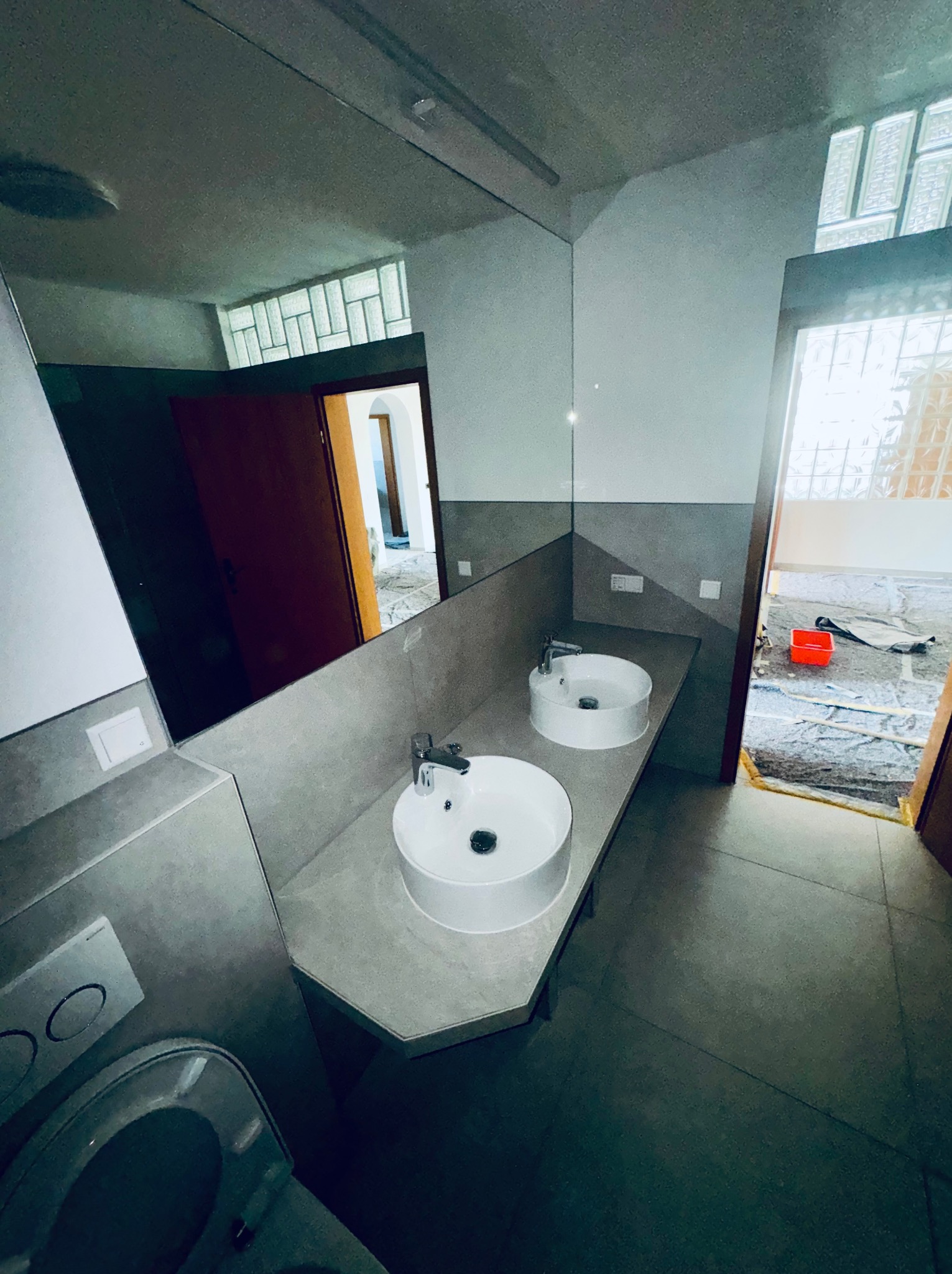 Nowoczesna łazienka z dwoma umywalkami na szarym blacie, duże lustro odbija widok na dalszą część domu. W tle widoczne okno z luksferami i prowizoryczne zabezpieczenia podłogi.
