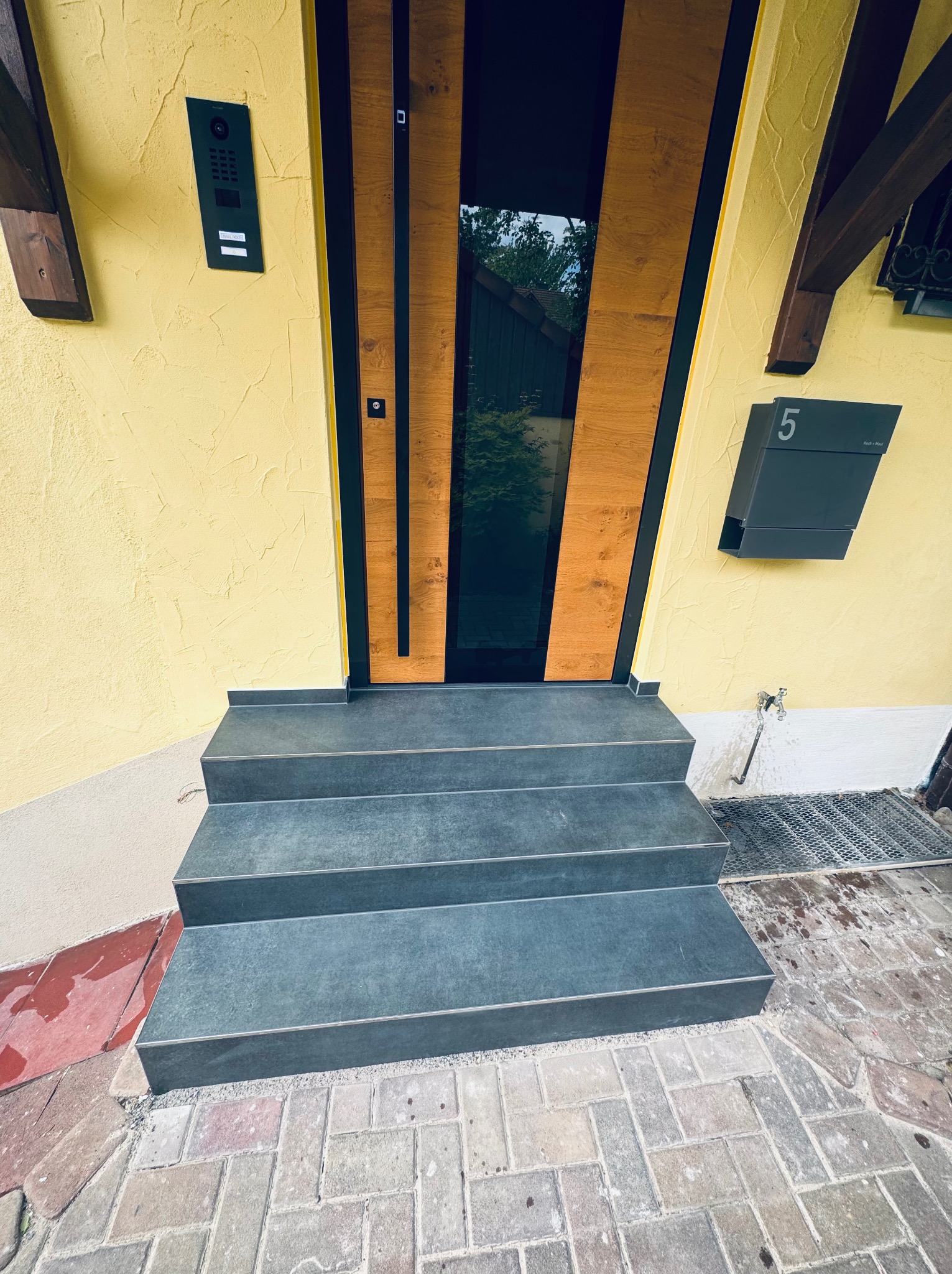 Nowoczesne wejście do domu w Bielawie: trzy stopnie z szarej płytki prowadzą do drzwi z drewnianymi panelami i czarnym szkłem. Żółta elewacja, skrzynka na listy nr 5.