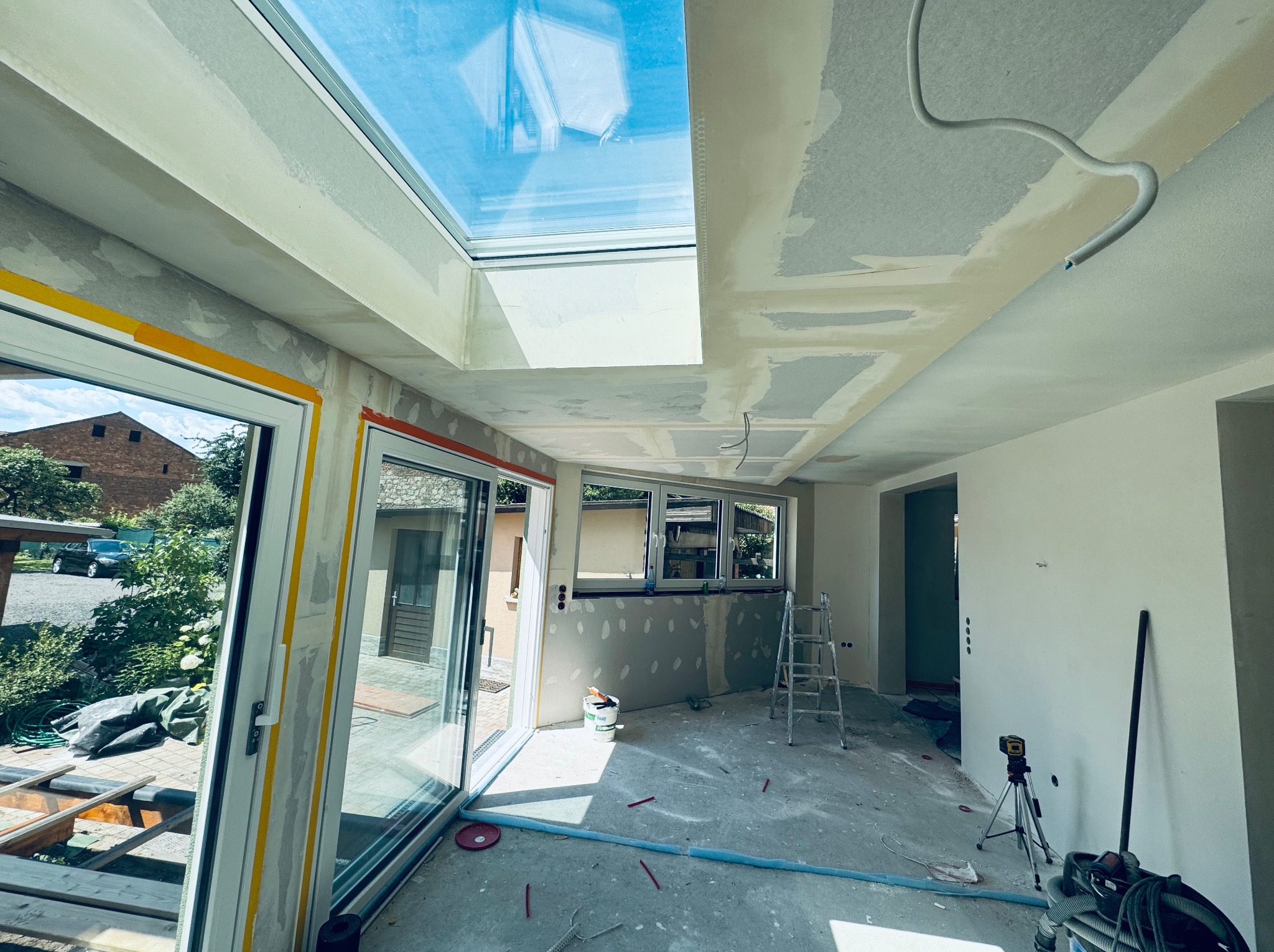 Wnętrze w trakcie wykańczania: okno dachowe, gładzie na suficie, widoczne narzędzia i drabina. Pomieszczenie z dużymi oknami i widokiem na zewnątrz.