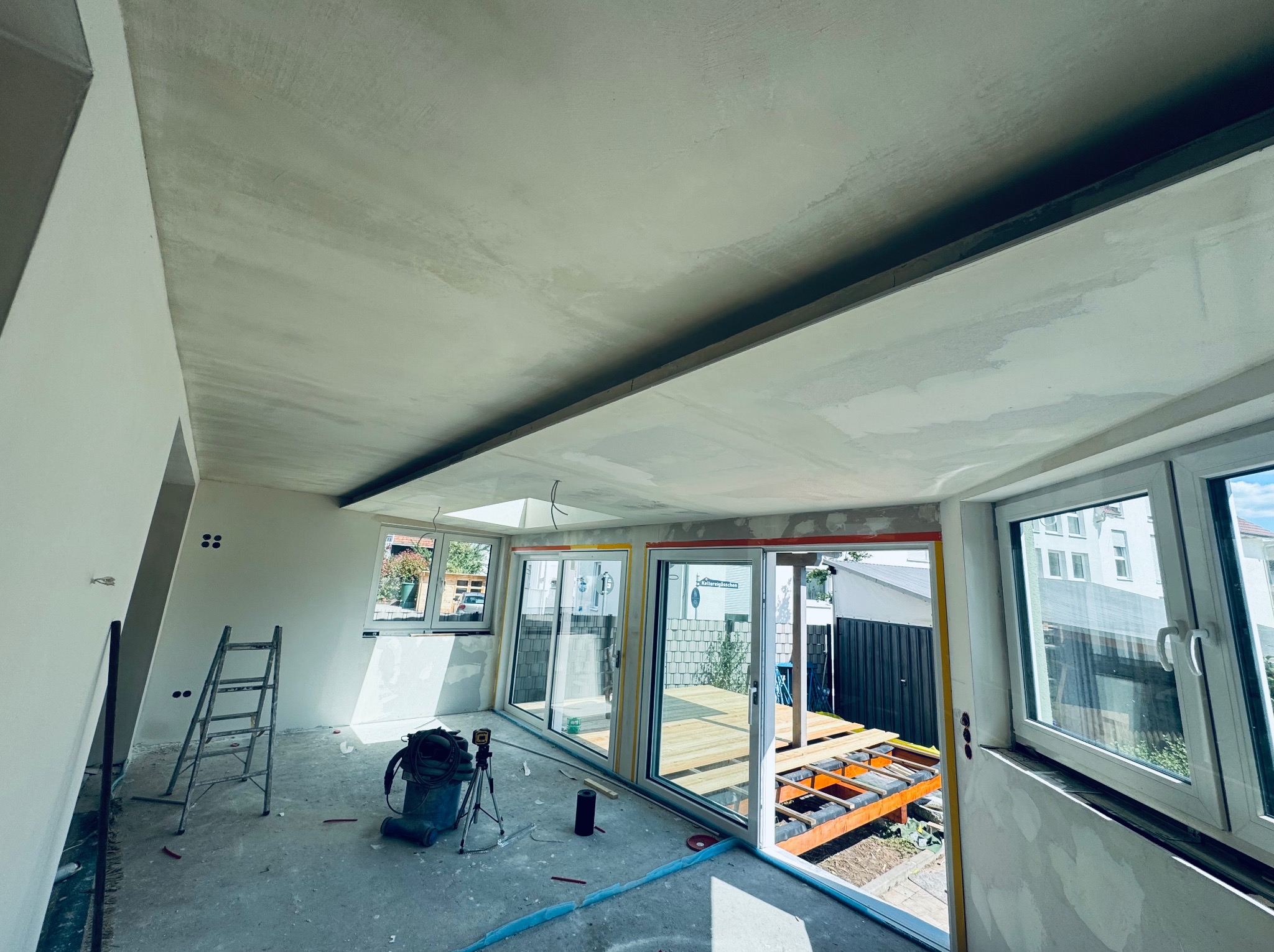 Wnętrze w trakcie wykańczania: gładzie na suficie, okna balkonowe z widokiem na taras, drabina i narzędzia. Widoczny etap prac wykończeniowych.